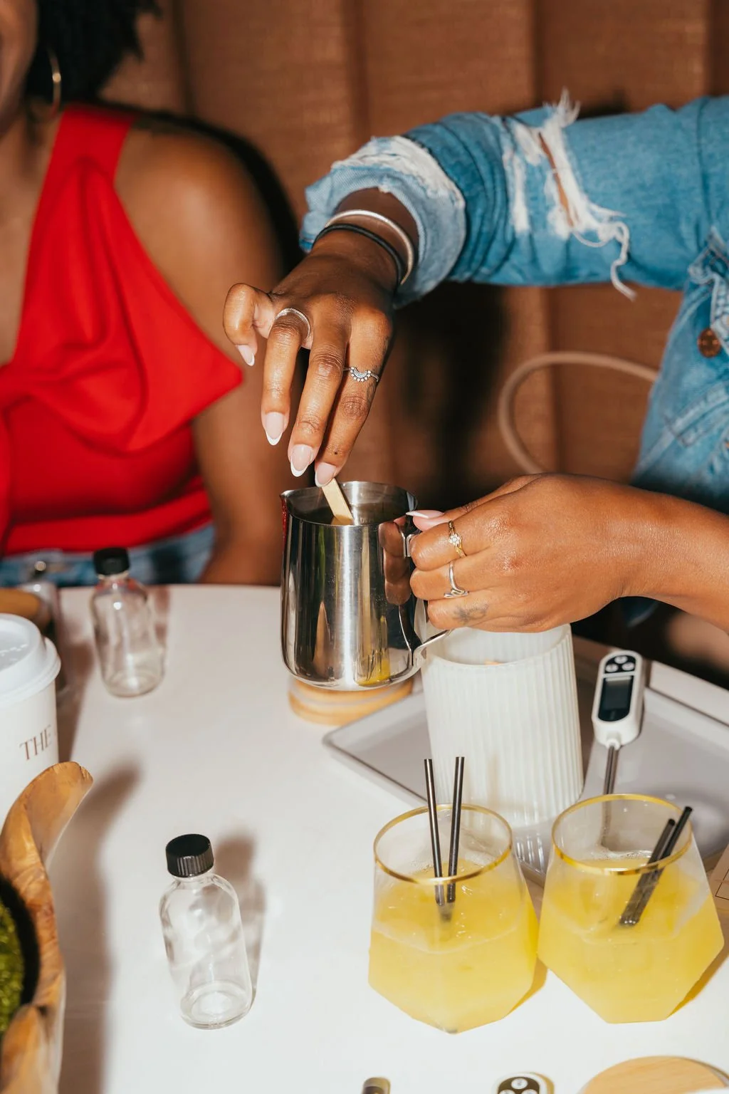 People preparing their own candles in a metal pitcher before pouring into their jars. There are also cocktail drinks on the table. 