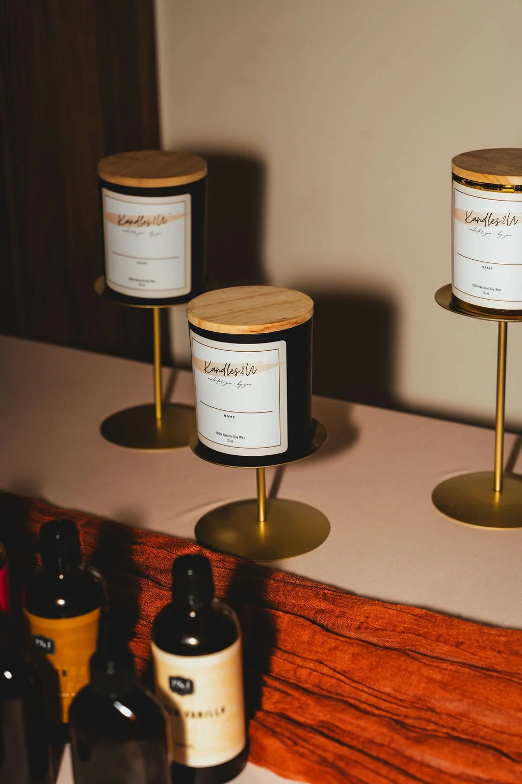 Three candles in black containers with wooden lids and white labels on gold stands, placed on a surface with bottles in the foreground. The candles are labeled 'Kandles2U' and are made of natural soy wax.