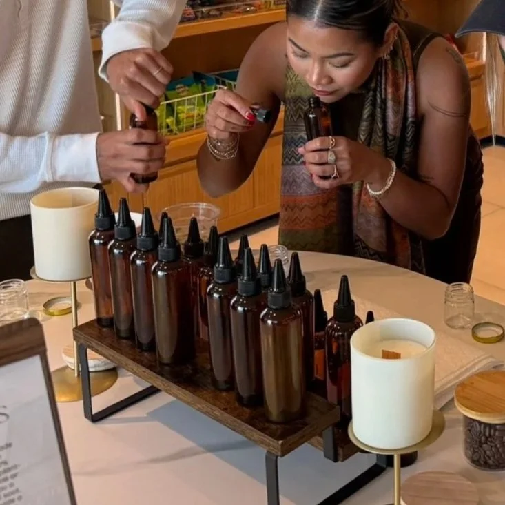 Two women at a table with amber glass dropper bottles, preparing essential oils or aromatic extracts, in a cozy indoor setting.