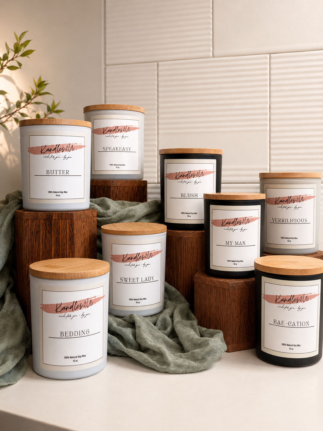 A collection of scented candles in white and black jars with wooden lids, each labeled with different names like Butter, Speakeasy, Blush, My Man, Sweet Lady, Bedding, Verrilicious, and Bae-cation, arranged on wooden blocks and draped fabric.