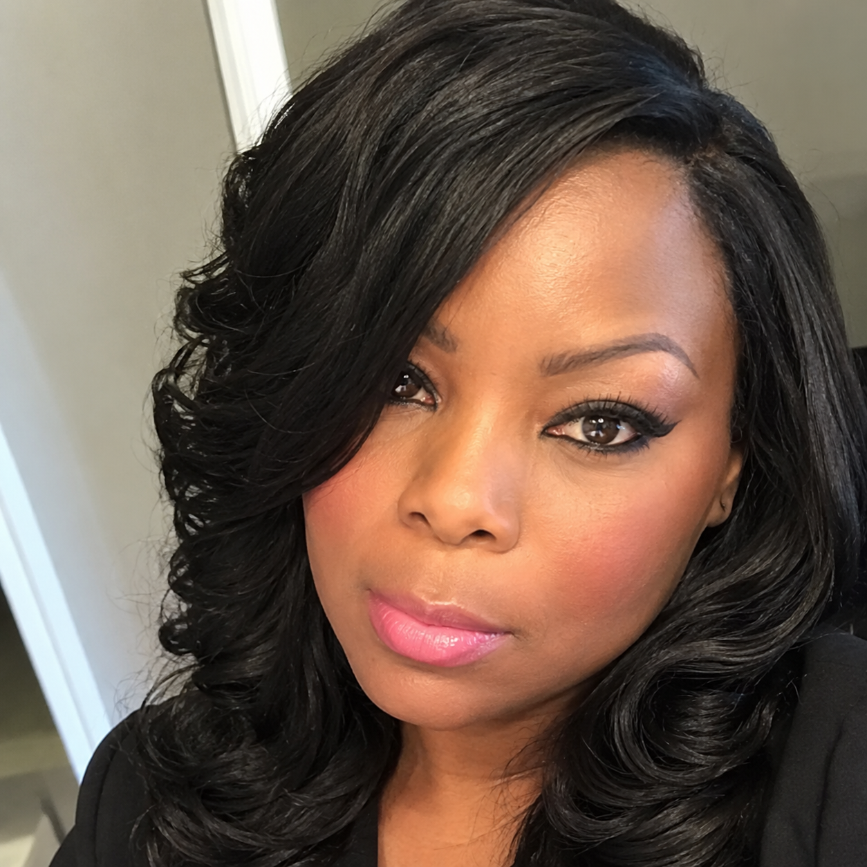 Close-up of a woman with long black wavy hair, makeup, pink lipstick, and wearing a black top, looking at the camera.