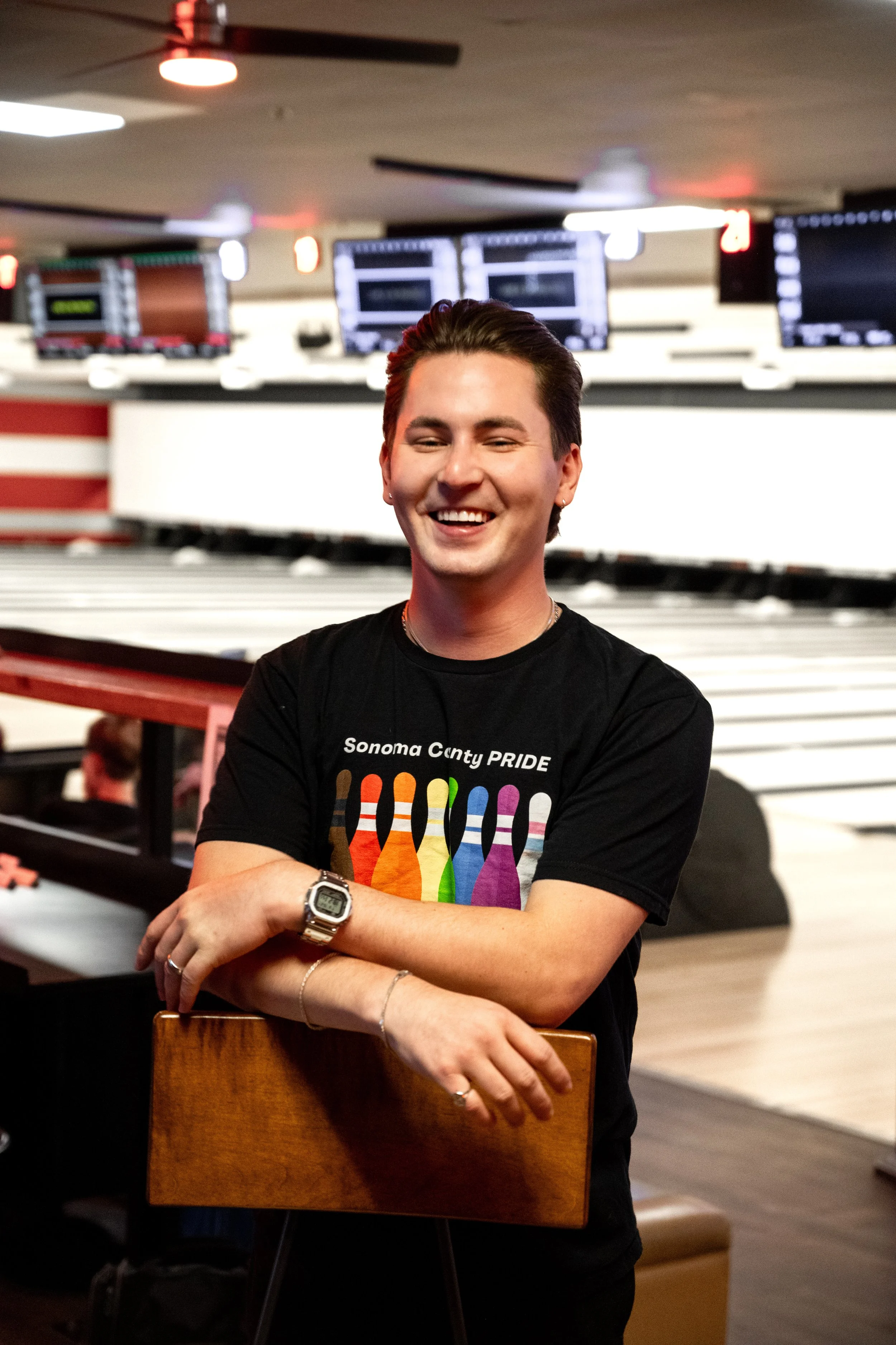 A slender man with a big smile and pushed back hair in a black Sonoma County Pride Bowling t-shirt, standing in front of lanes in a bowling alley