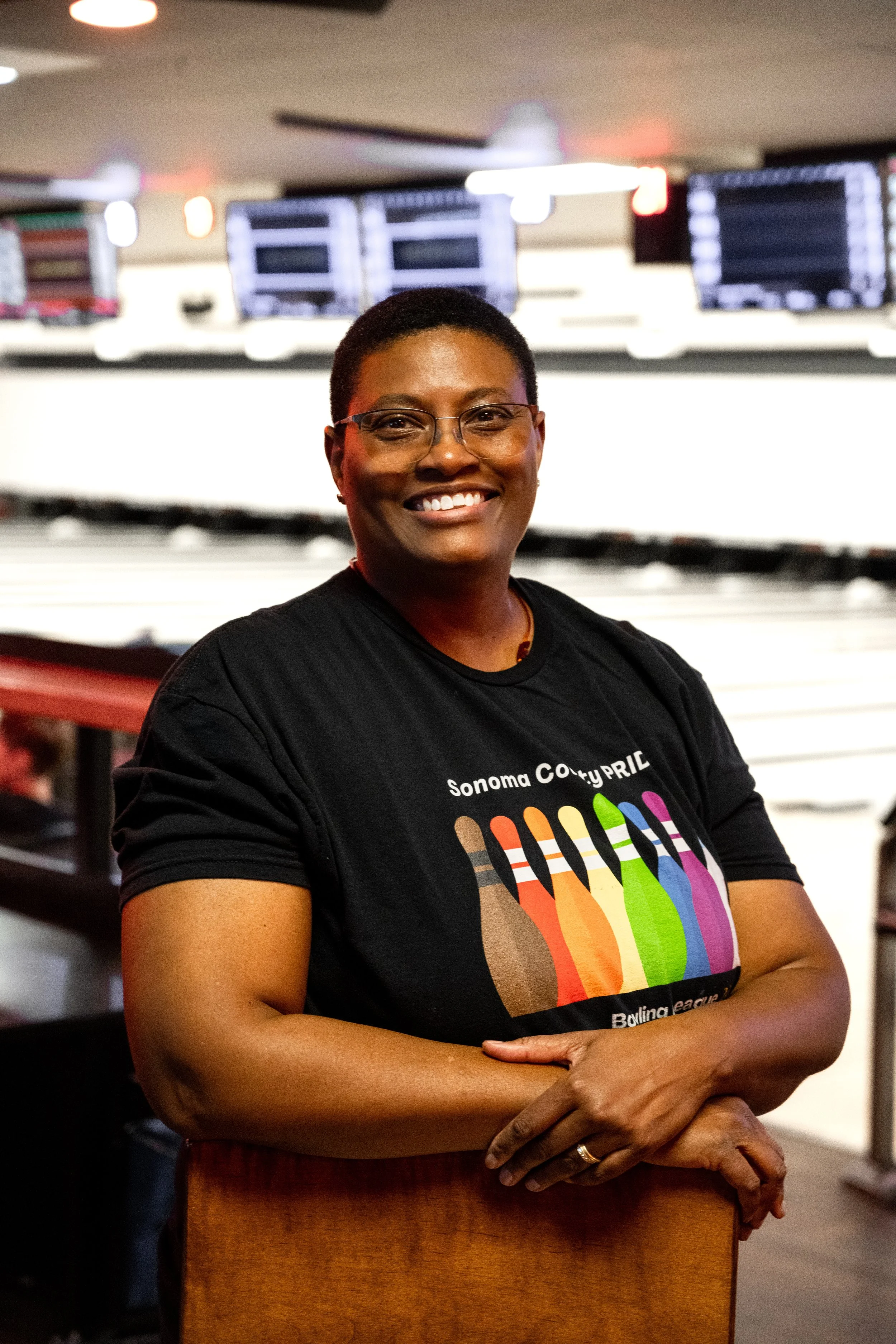 A smiling woman with thin glasses in a black Sonoma County Pride Bowling t-shirt, standing in front of lanes in a bowling alley