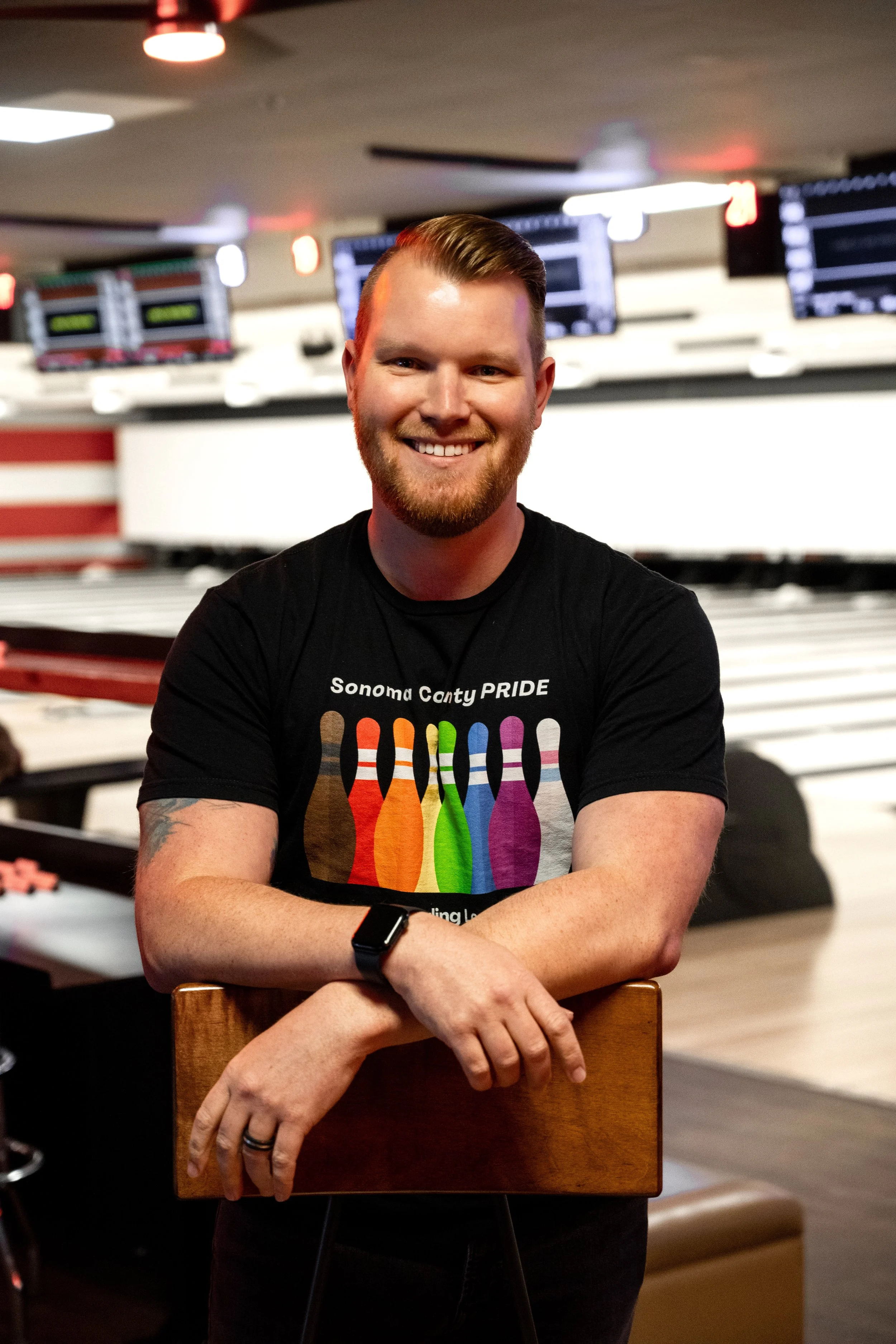A tall smiling man with blonde hair and arms crossed in a black Sonoma County Pride Bowling t-shirt, standing in front of lanes in a bowling alley