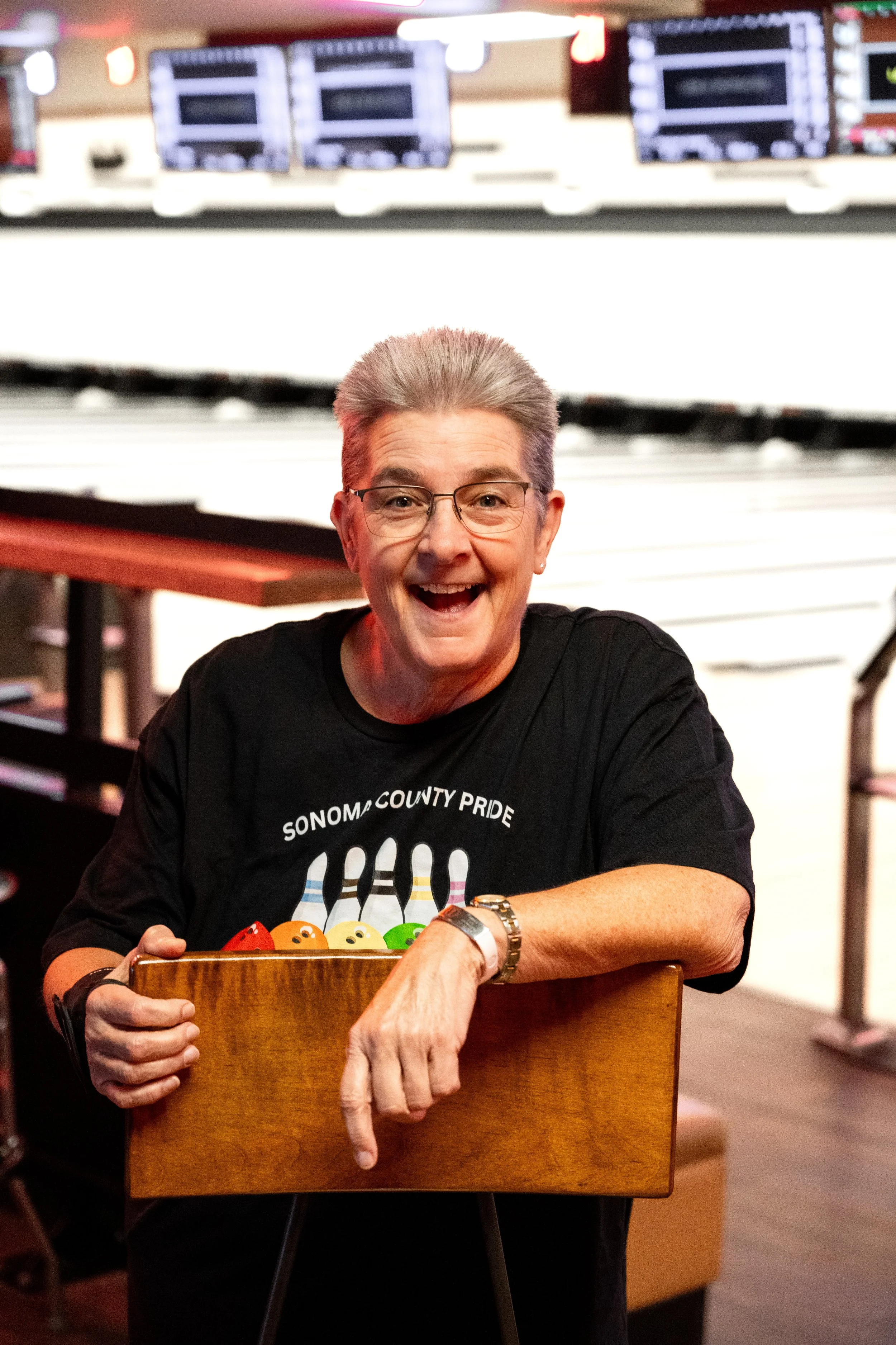 A joyful woman with glasses and spiked gray hair in a black Sonoma County Pride Bowling t-shirt, standing in front of lanes in a bowling alley