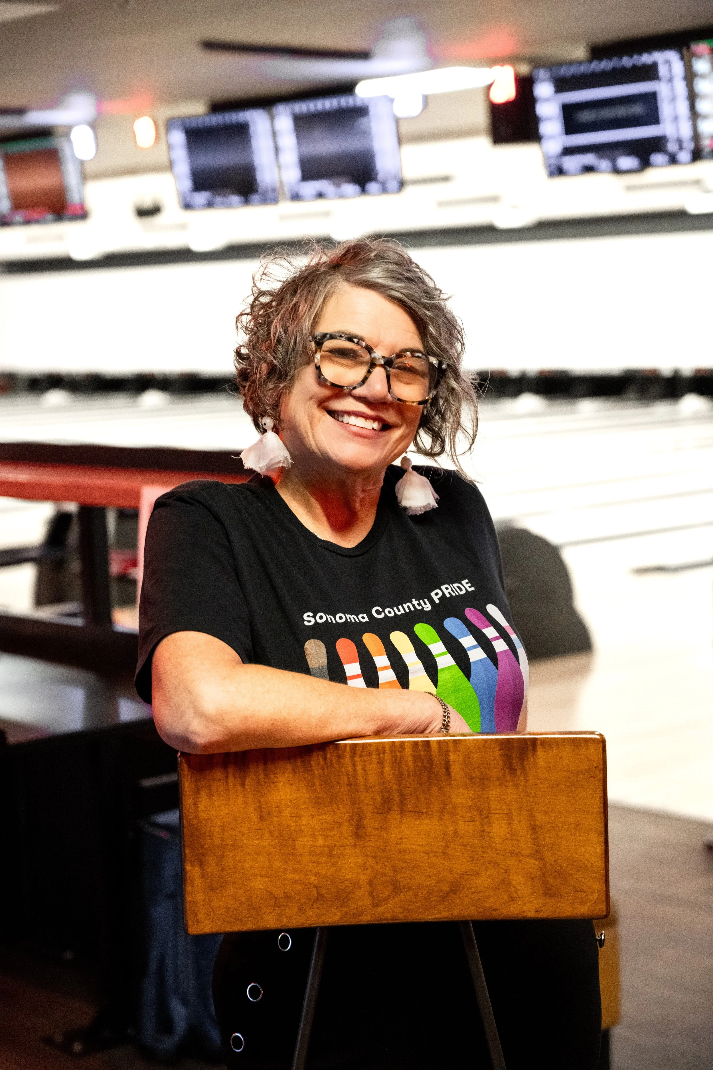A fun woman with dangle earrings and short messy hair in a black Sonoma County Pride Bowling t-shirt, standing in front of lanes in a bowling alley