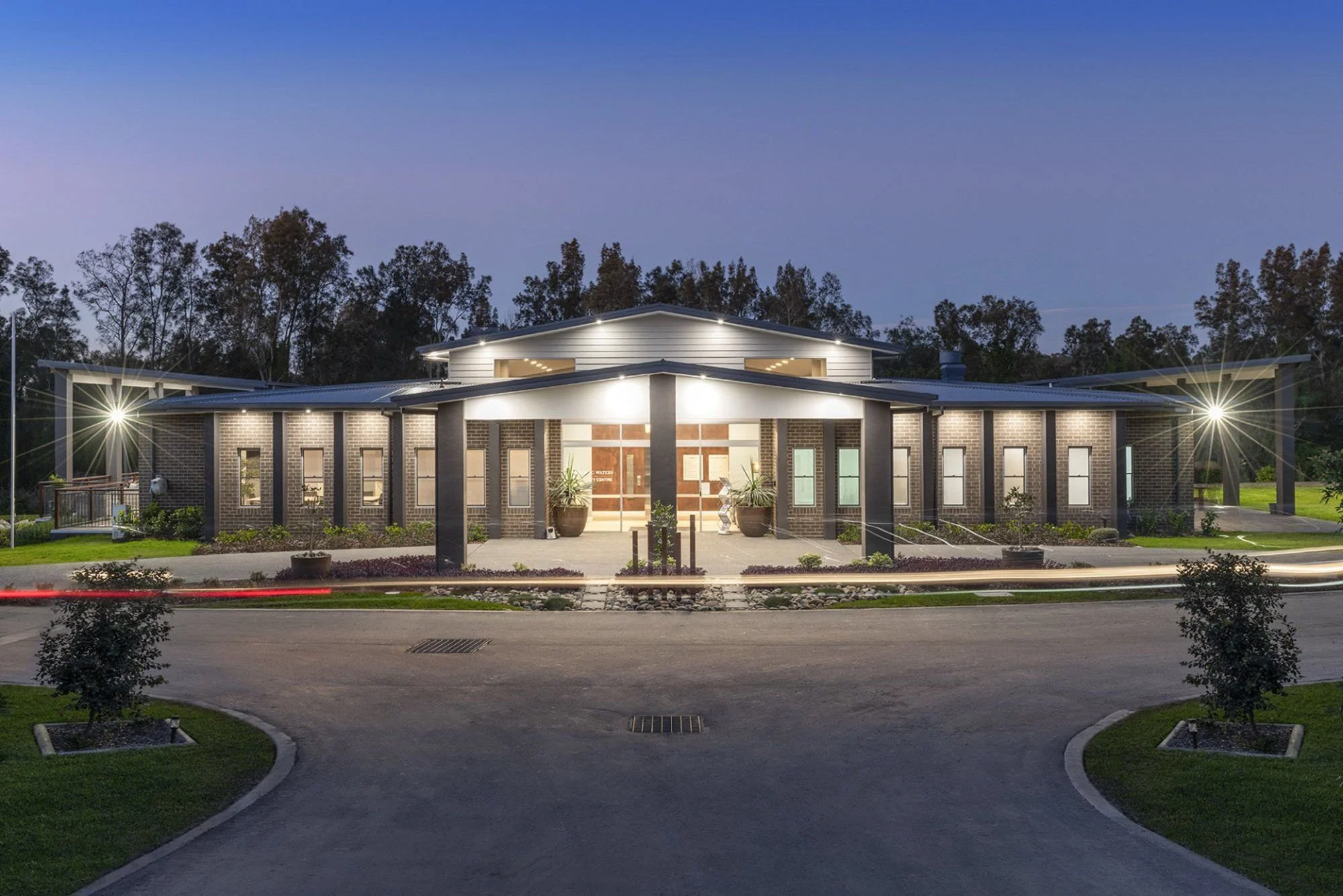 A modern brick building with large windows, illuminated at dusk, surrounded by landscaped greenery and a circular driveway.