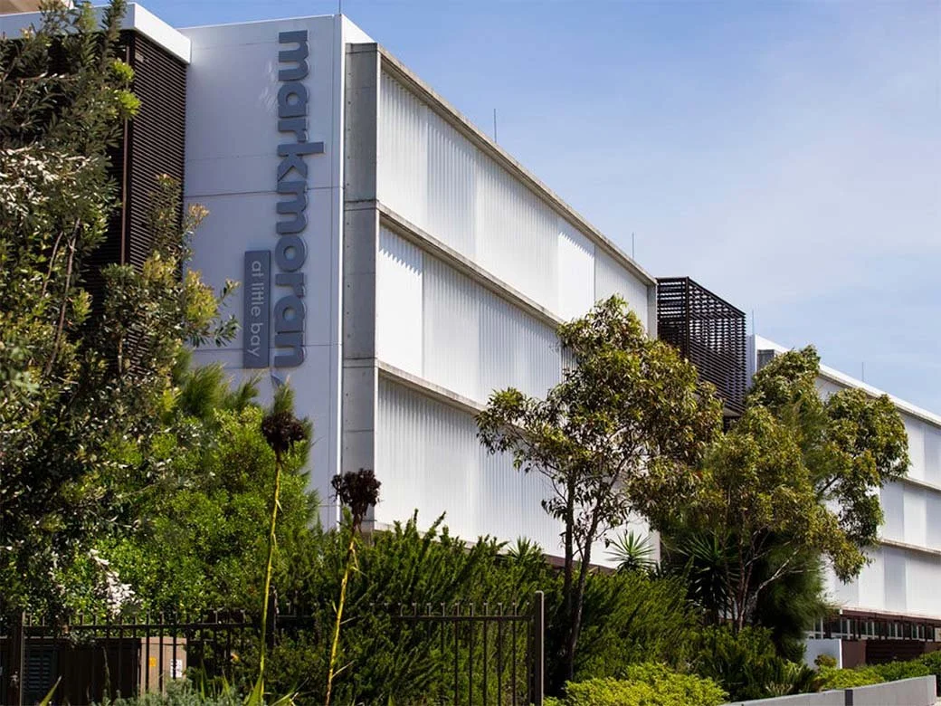 Modern building with blue sign reading "marlborough art the body" on a light gray facade, surrounded by lush green trees and shrubs, under a partly cloudy sky.
