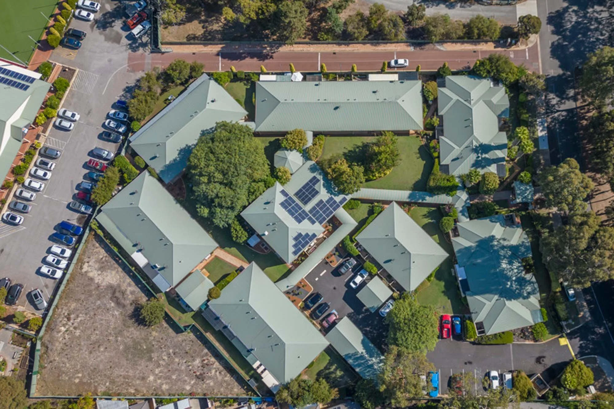 Aerial view of a residential complex with multiple buildings, green lawns, trees, parking lots, and solar panels on some rooftops.