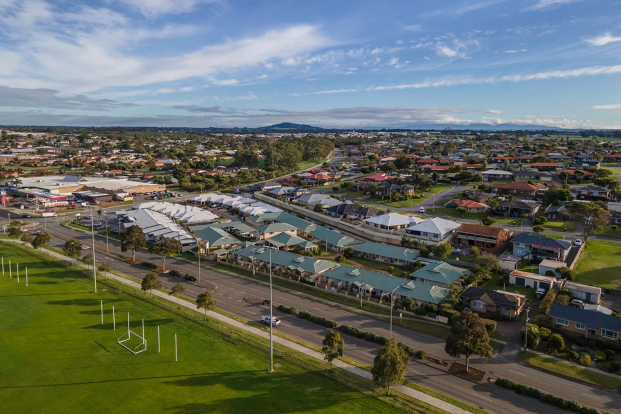 Aerial view of a suburban residential area with houses, a small shopping center with a parking lot, and a sports field with goal posts, under a partly cloudy sky.