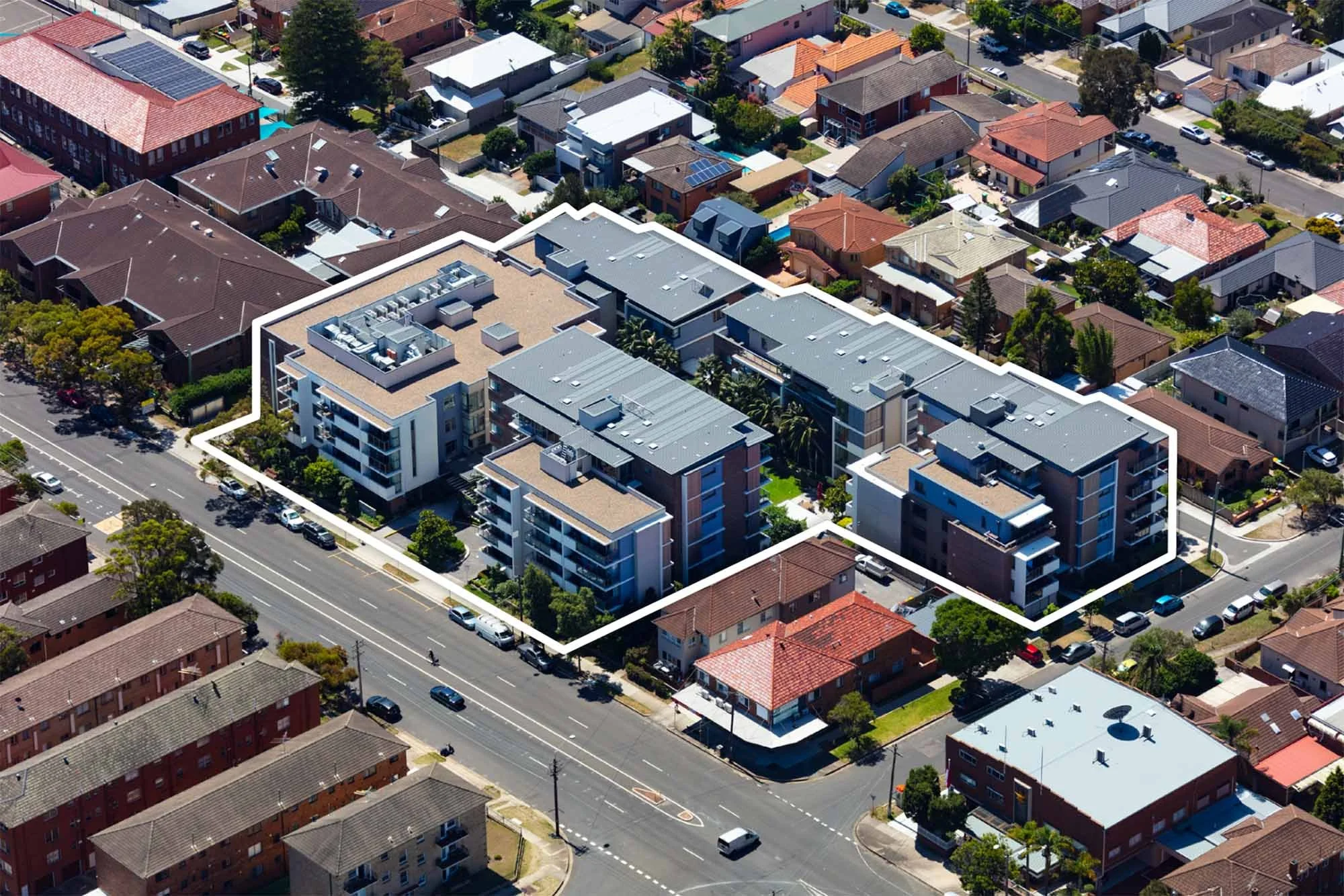 An aerial view of a residential area featuring a modern apartment complex outlined in white, surrounded by single-family homes and streets with cars and trees.