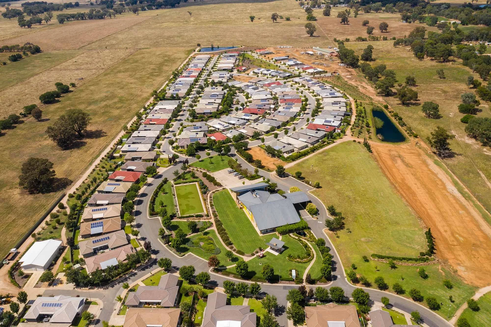 Aerial view of a suburban neighborhood with winding roads, houses, a central park with a pond, and surrounding open fields.