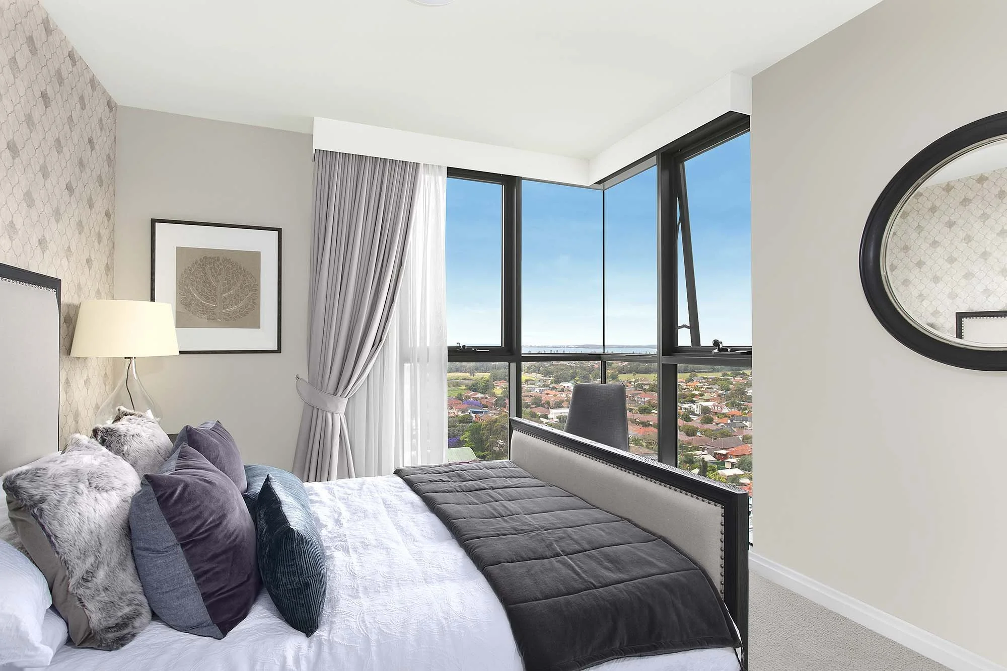 Bedroom with large windows, bed with multiple pillows, a lamp, framed artwork, a round mirror, and city view outside.
