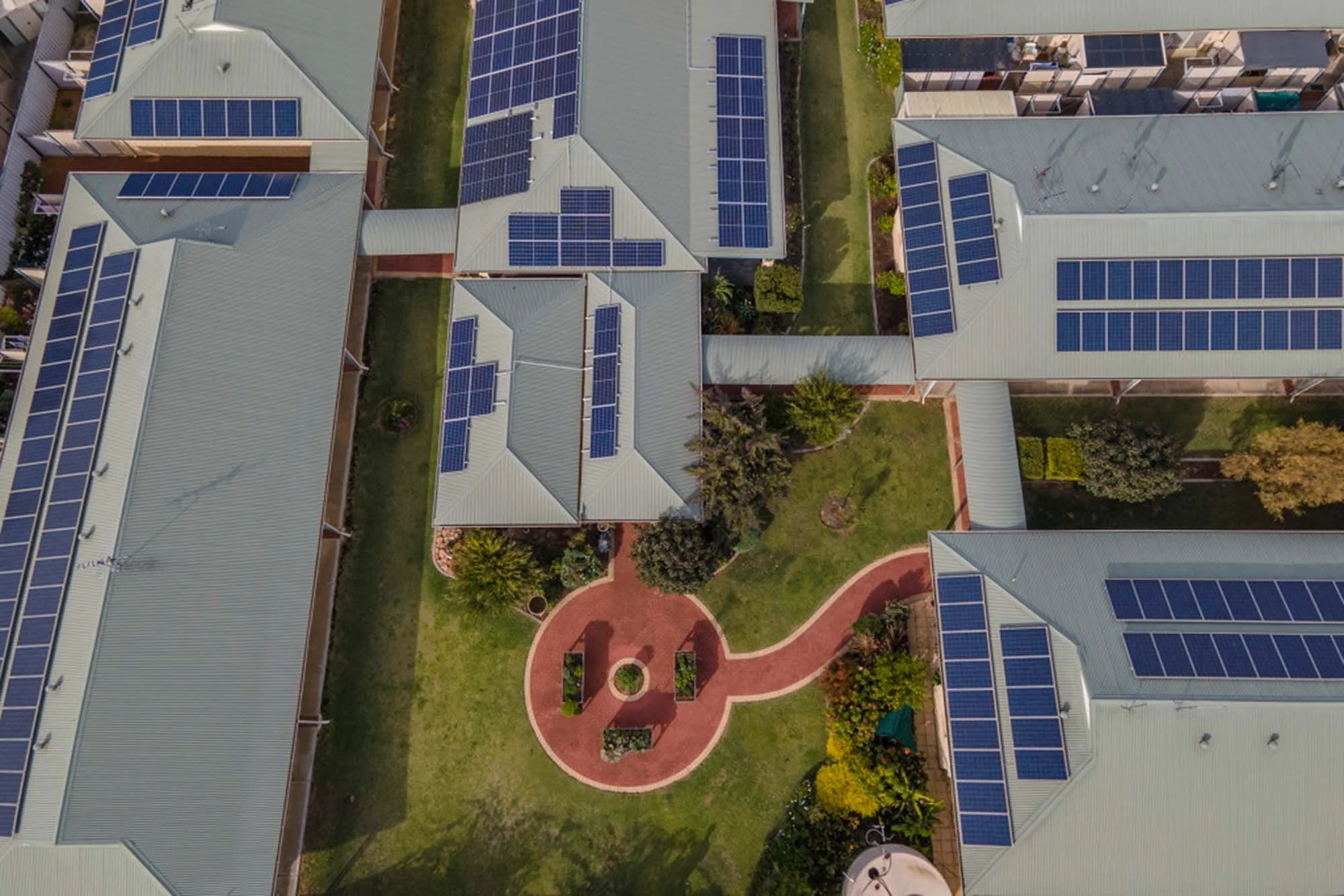 Aerial view of residential complex with multiple buildings featuring solar panels on metal roofs and a green courtyard with a circular brick pathway and landscaped garden.