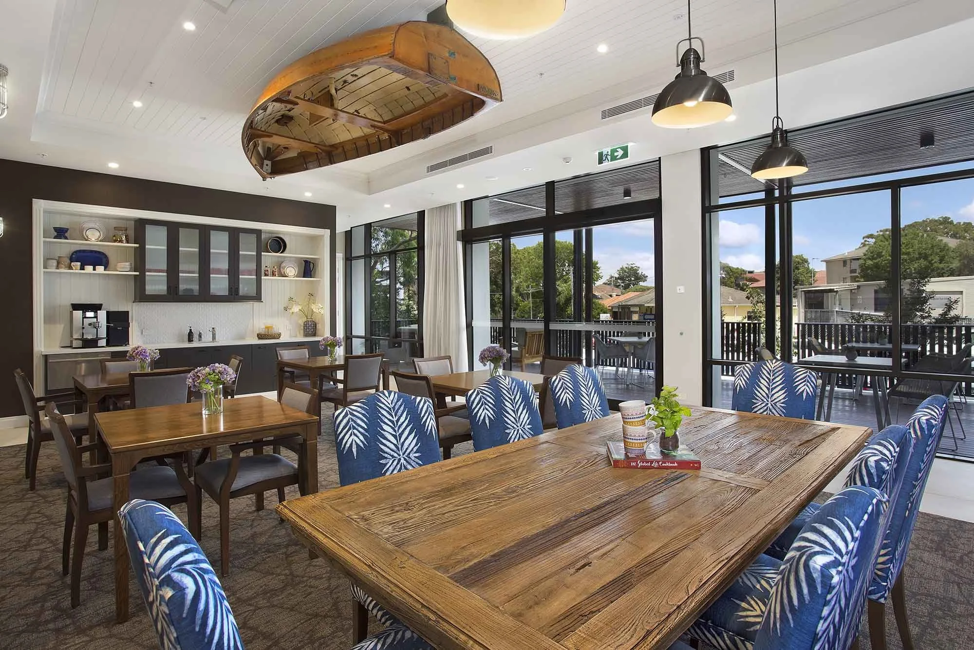 A bright, modern dining area with wooden tables and blue chairs with white leaf patterns, large windows showing an outdoor balcony and trees, and a wall with shelves and a coffee station.