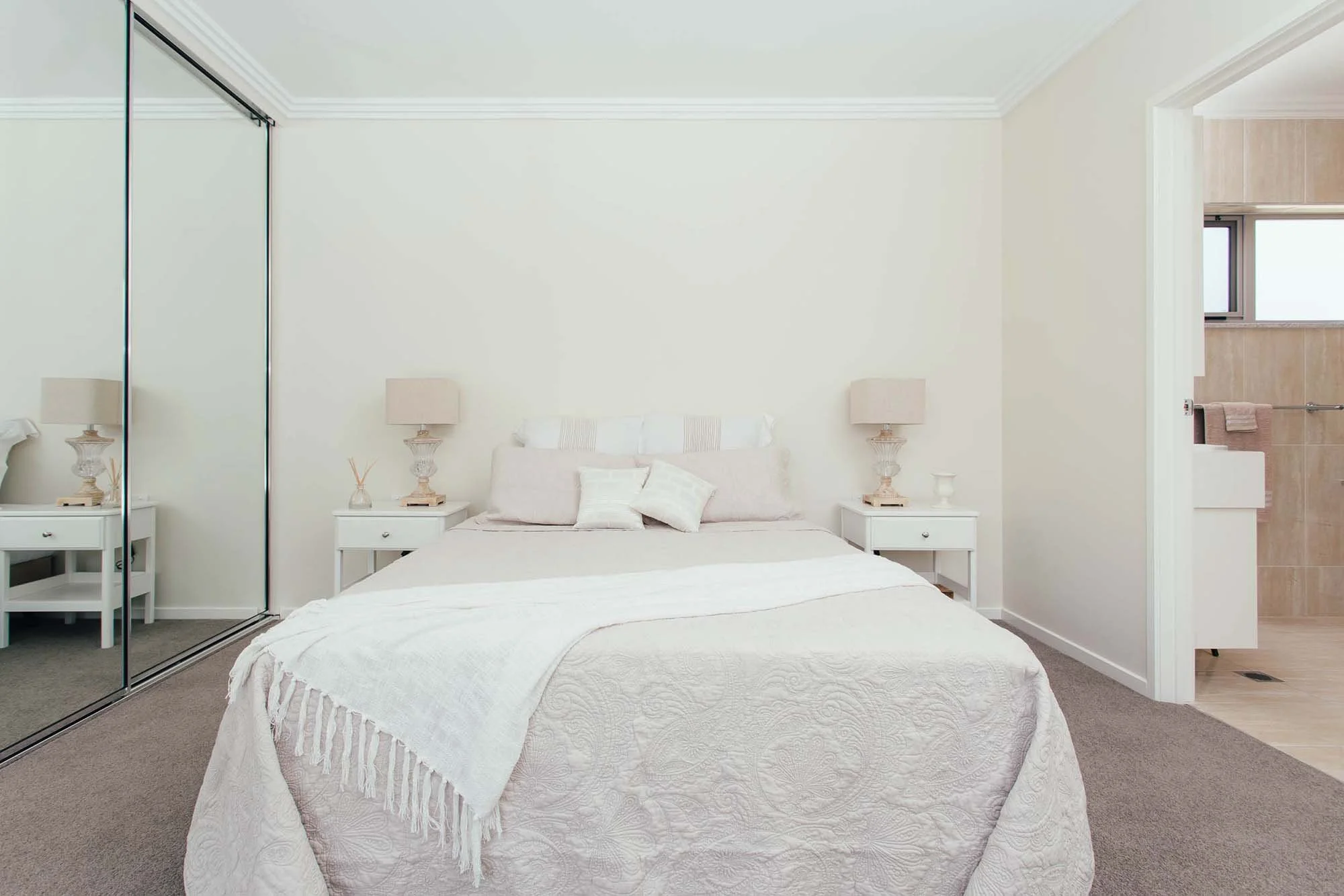 A bedroom with a white bed, beige pillows, white bedside tables with lamps, a mirrored closet, and a doorway leading to a bathroom with beige tiles and a window.