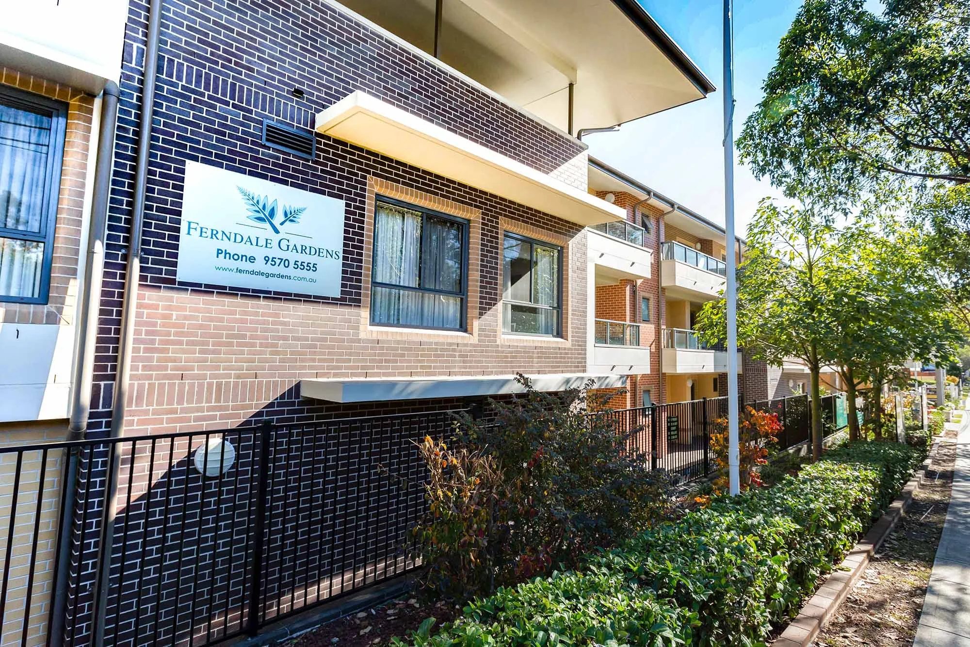 Exterior view of a residential apartment building with a Ferndale Gardens sign, brick walls, balconies, and lush greenery in front.