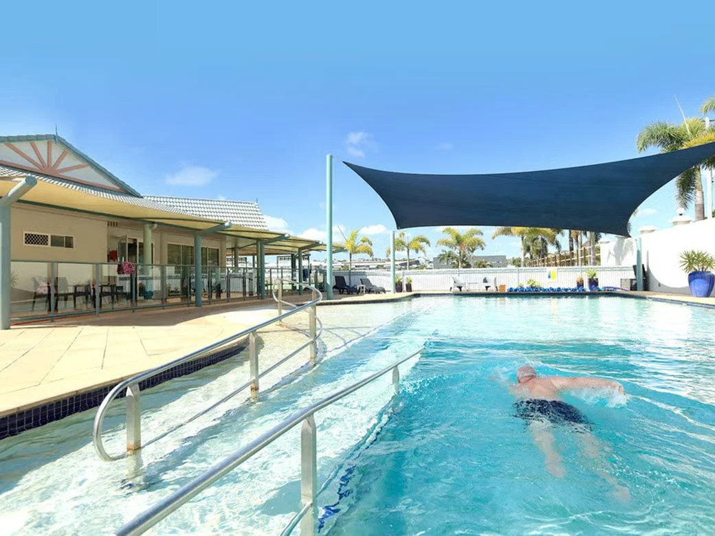 Swimming pool with a man swimming, shaded area with sail shade, building with porch, and palm trees in the background under a clear blue sky.