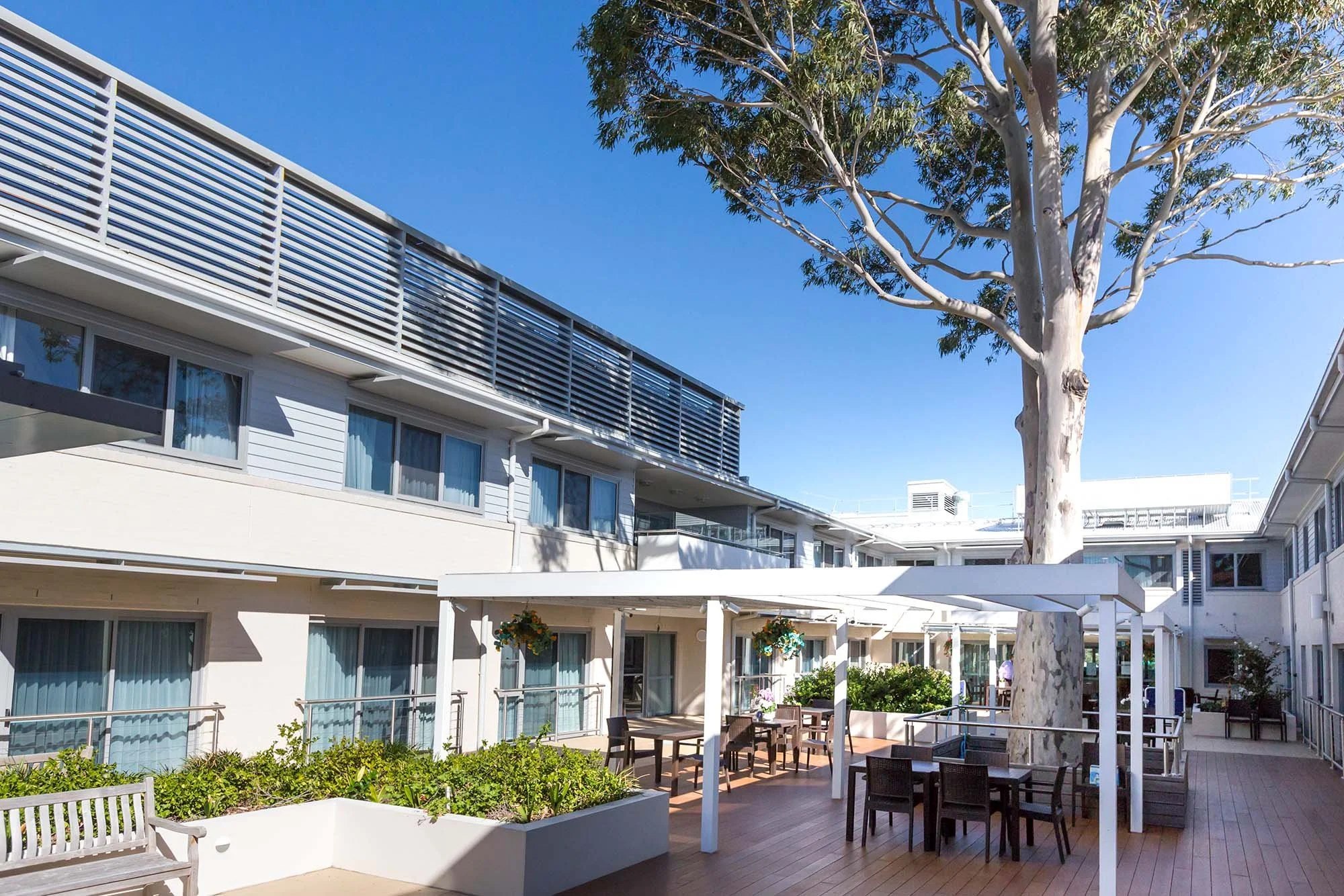 Modern outdoor patio with tables and chairs, green plants, large tree, multi-story residential building, and clear blue sky.