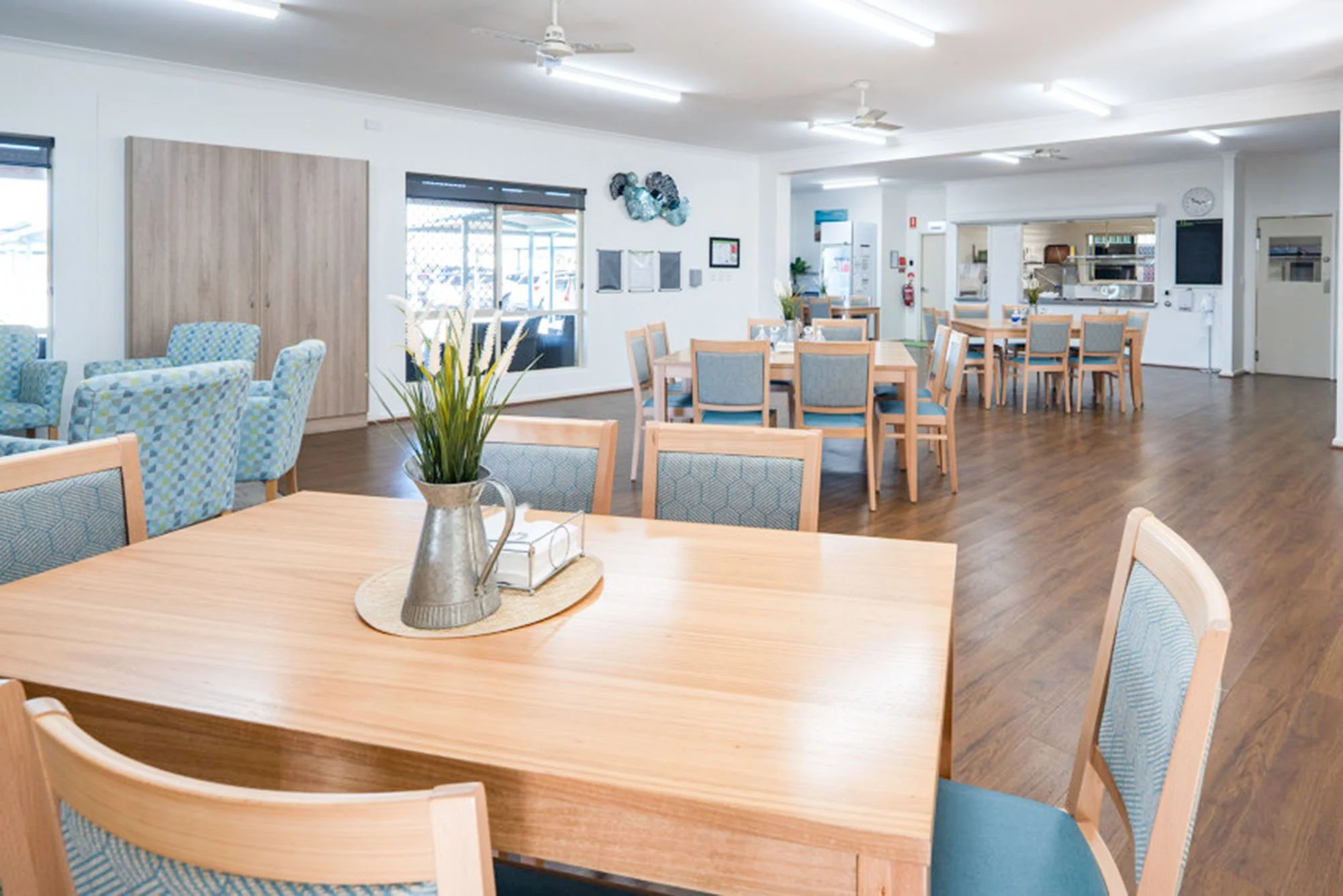 Empty dining area with wooden tables, chairs with blue upholstery, and a centerpiece with flowers in a pitcher.
