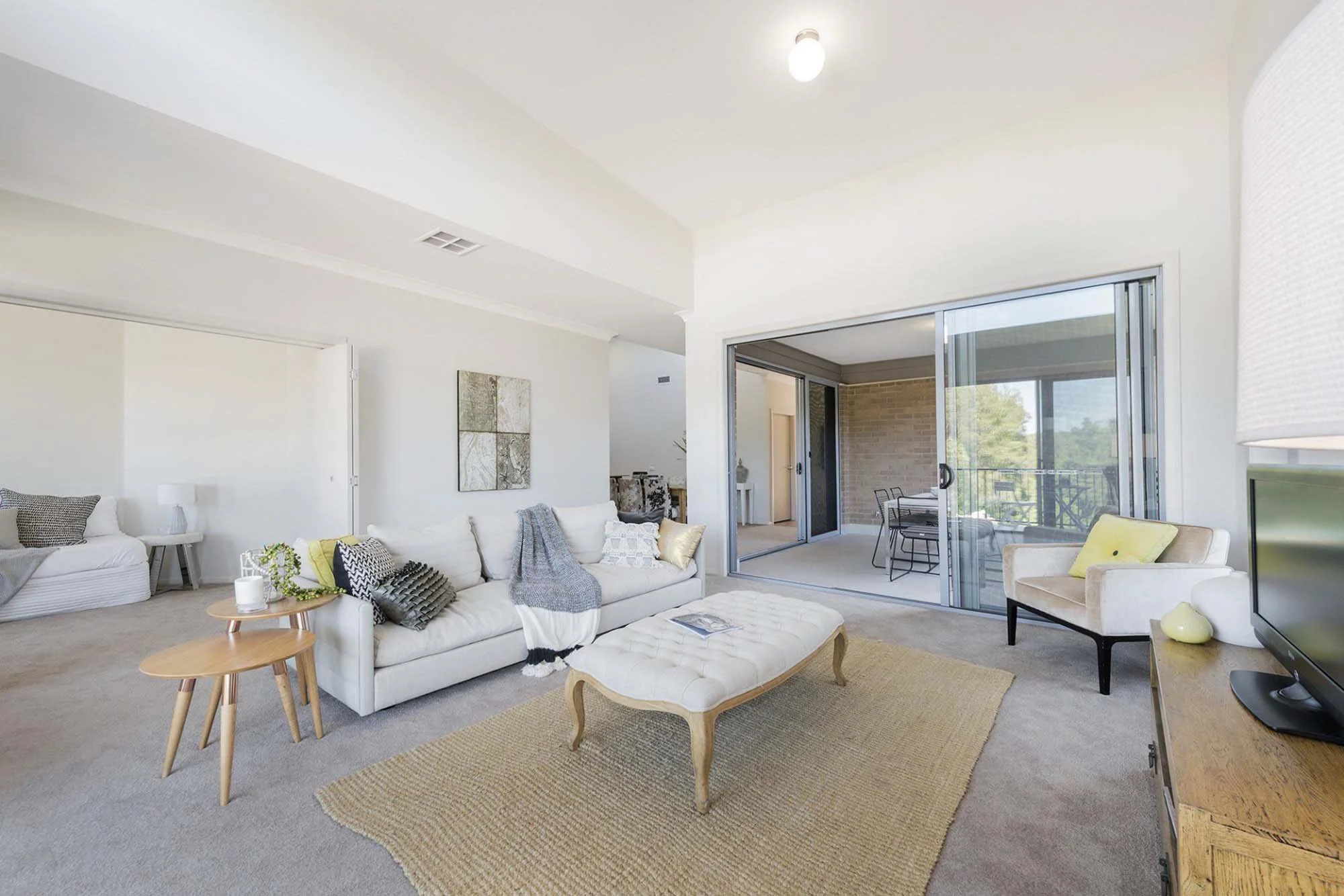 Living room with beige carpet, white sofa with decorative pillows, small wooden coffee table, beige armchair, TV on wooden stand, sliding glass door leading to balcony with a table and chairs, and an outdoor view of trees.