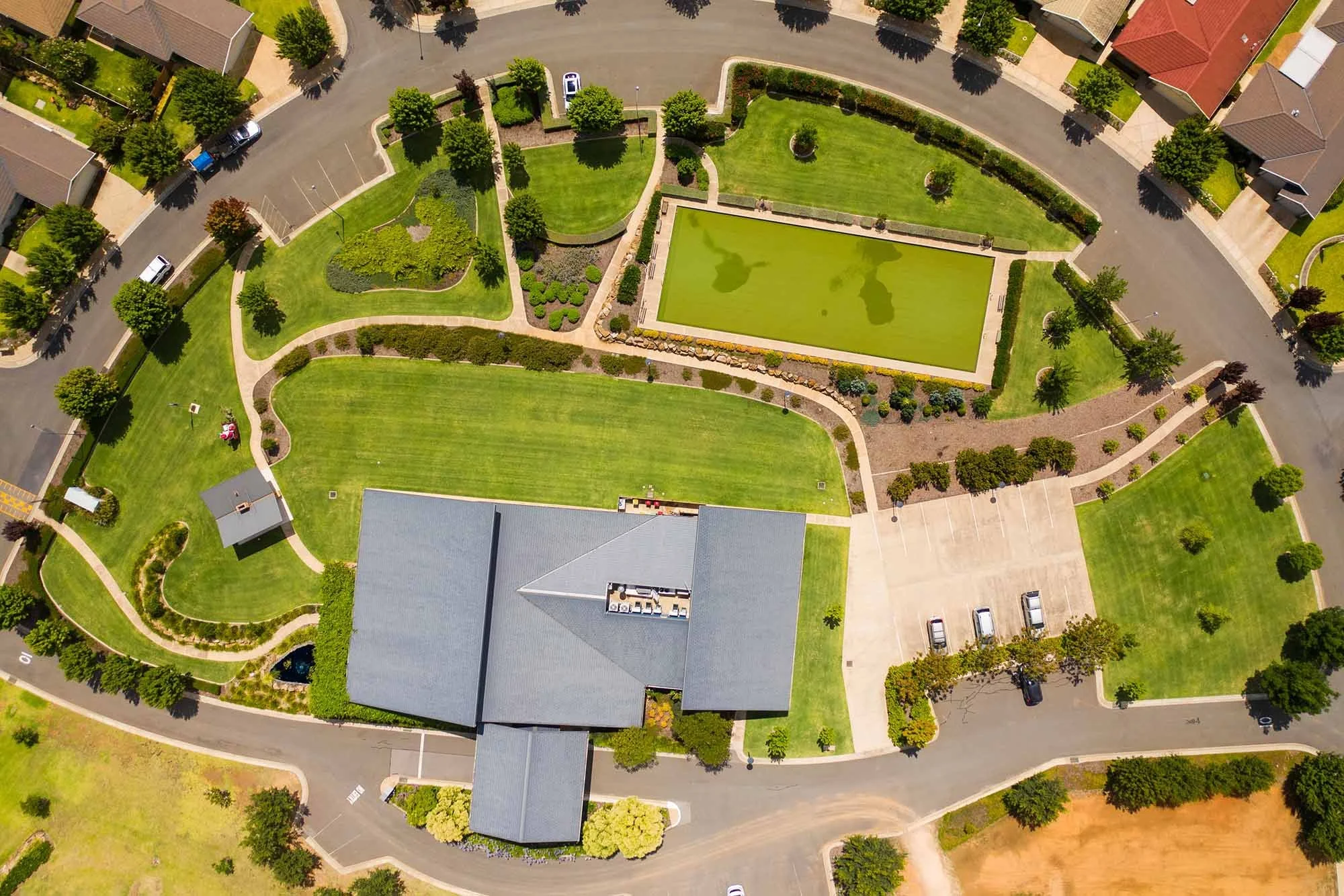 An aerial view of a residential community park with walking paths, green lawns, trees, a pond, and parking lot surrounded by houses.