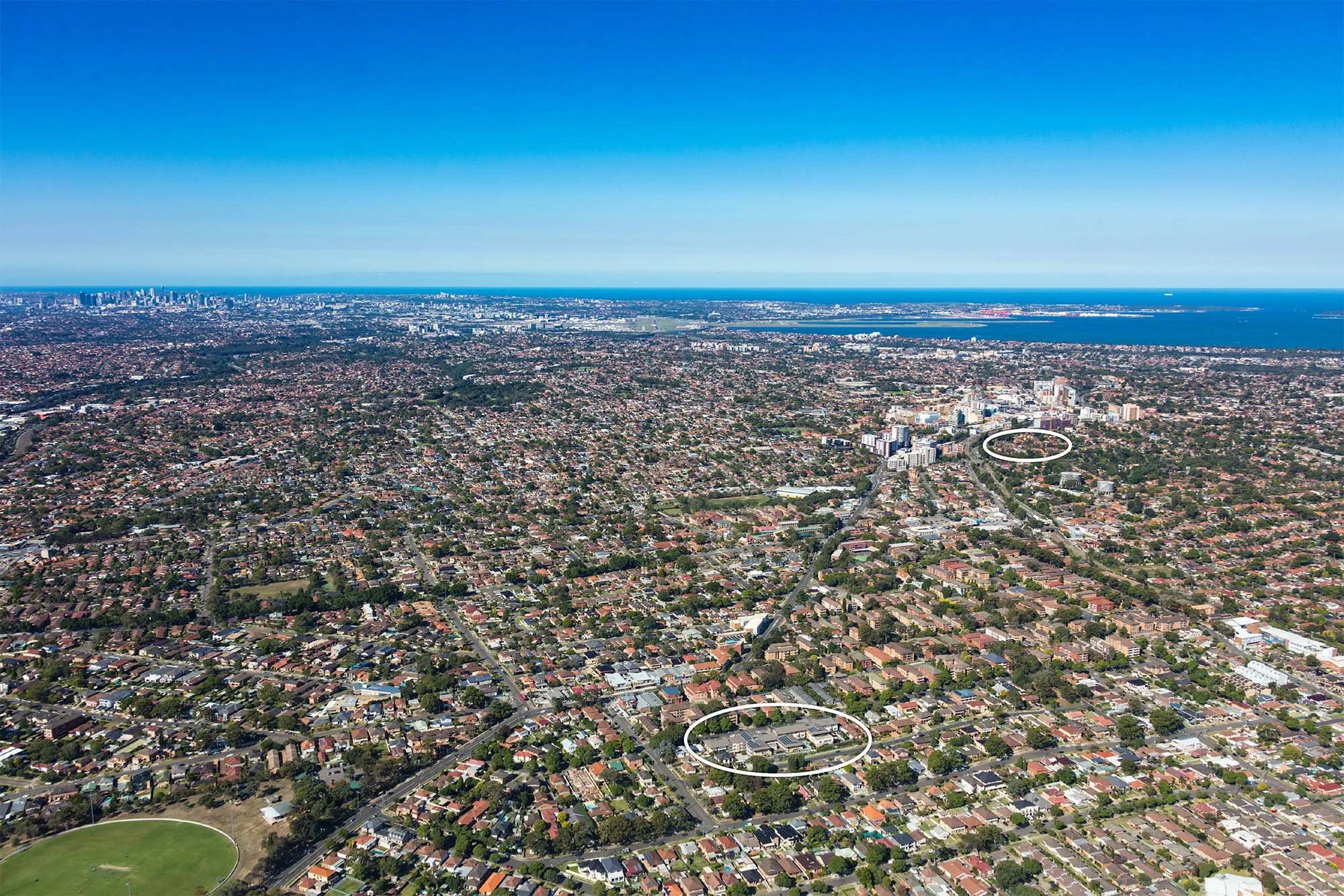 Aerial view of a city with residential neighborhoods, commercial buildings, a sports field, and a distant shoreline with high-rise buildings