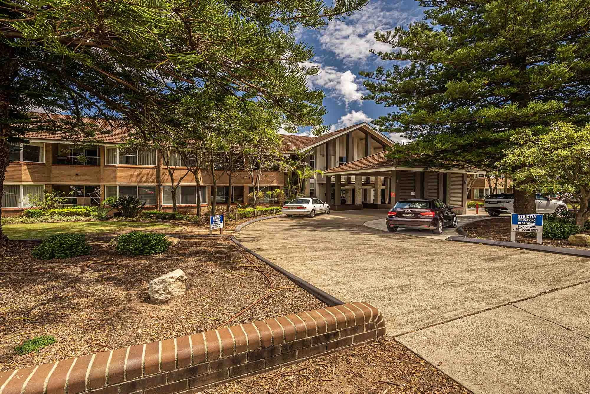 A hotel or motel building with a driveway in front, surrounded by trees and greenery, with several parked cars near the entrance.