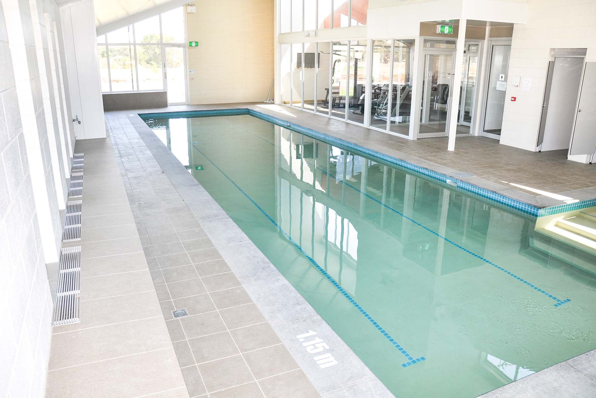 Indoor swimming pool with clear water, tiled floor, and fitness area visible through glass doors in the background.