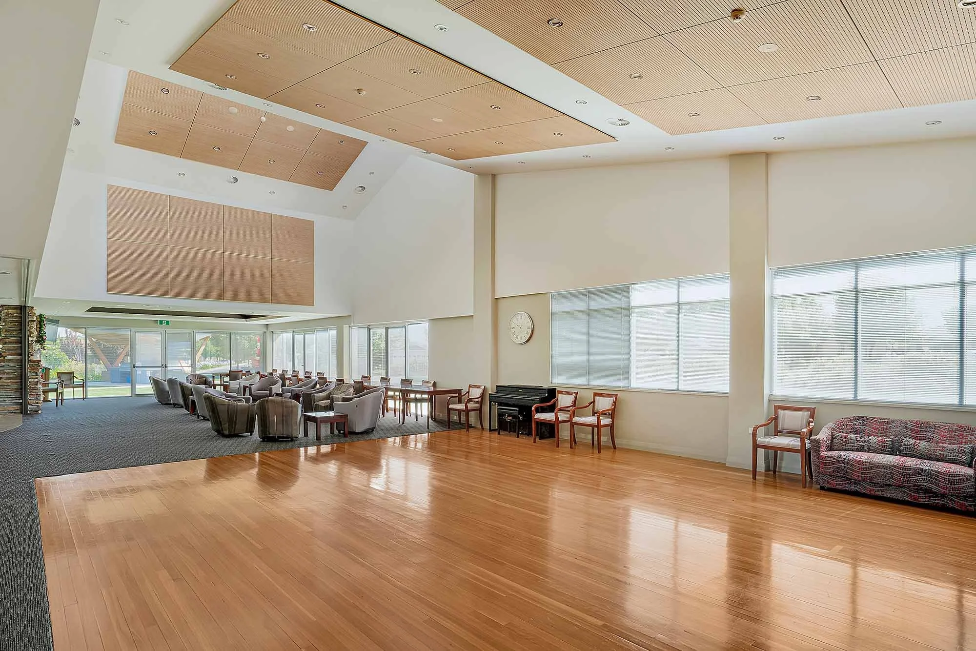 Spacious room with wooden flooring, large windows with blinds, and furniture including chairs, a sofa, and a piano.