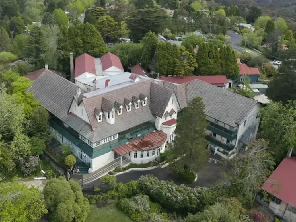 Aerial view of a large, multi-story house surrounded by trees and greenery, with several smaller buildings and houses nearby.