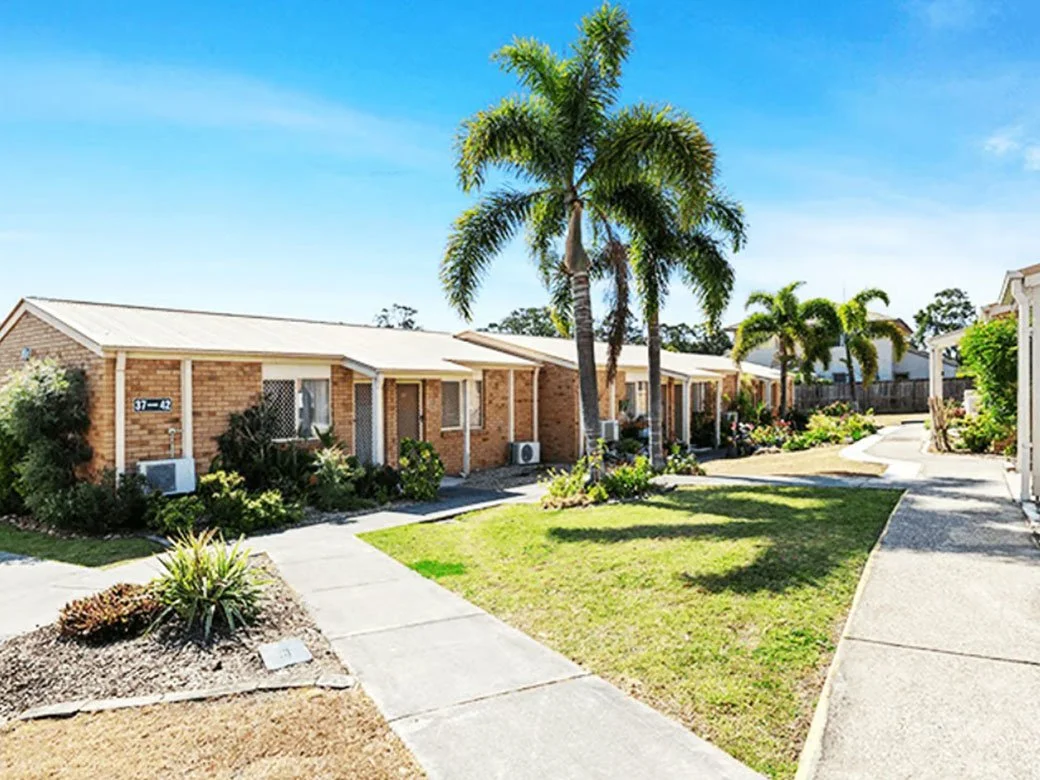 A row of single-story brick apartments with small front yards, tall palm trees, and a bright blue sky.