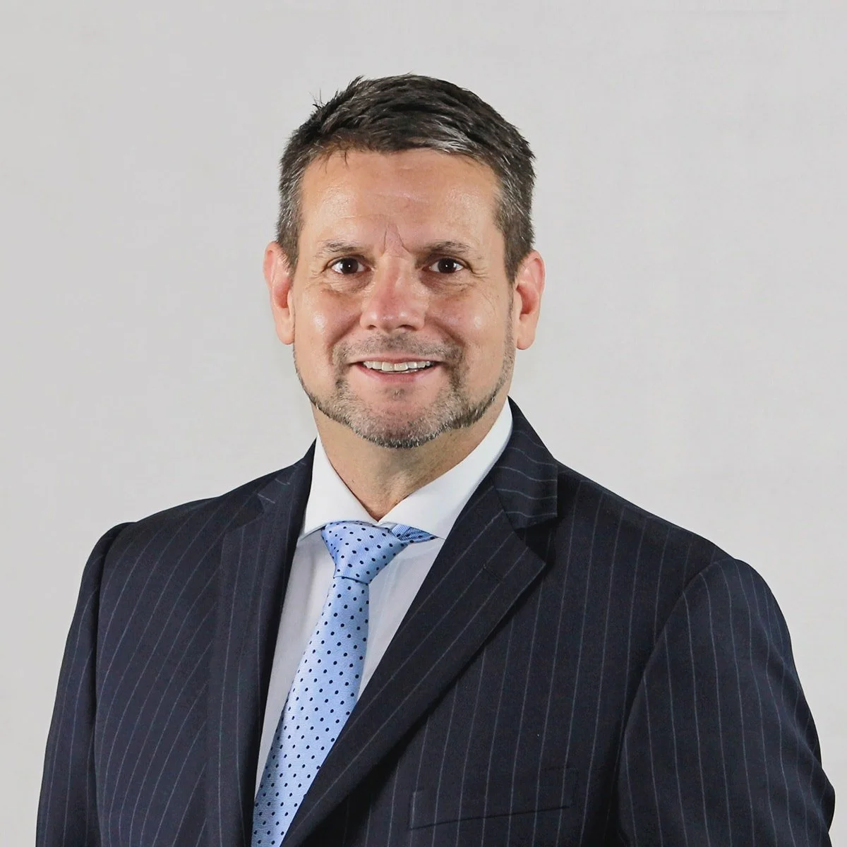 A middle-aged man with dark hair, a beard, and a mustache, wearing a dark pinstripe suit, white shirt, and a light blue polka dot tie, smiling against a plain light gray background.