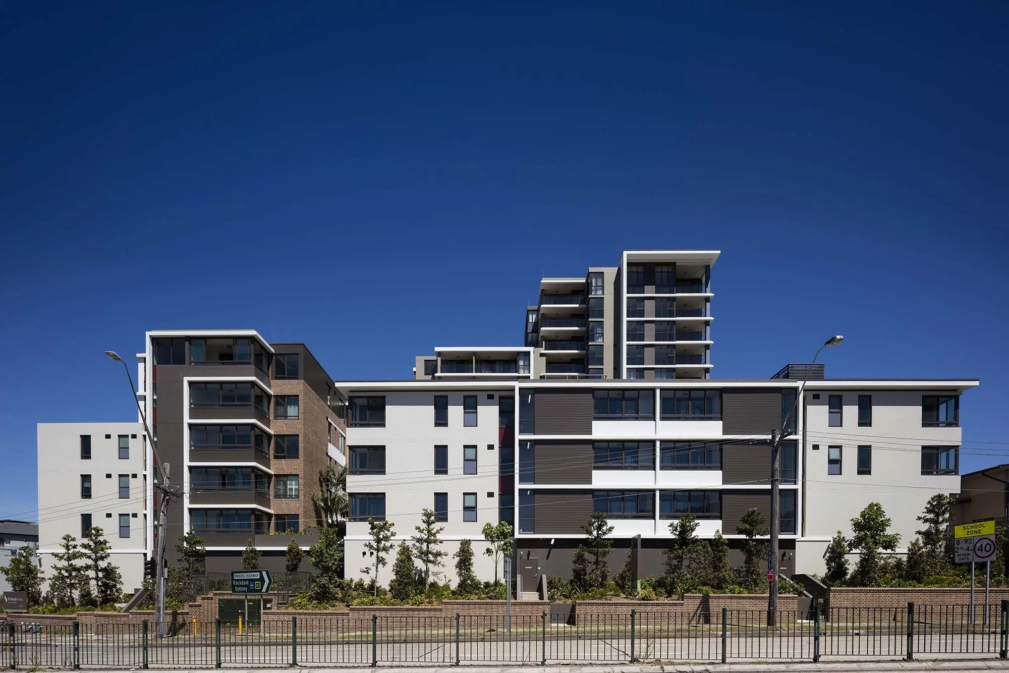 Modern multi-story apartment buildings with white, gray, and brown exteriors, surrounded by small trees and street lights under a clear blue sky.