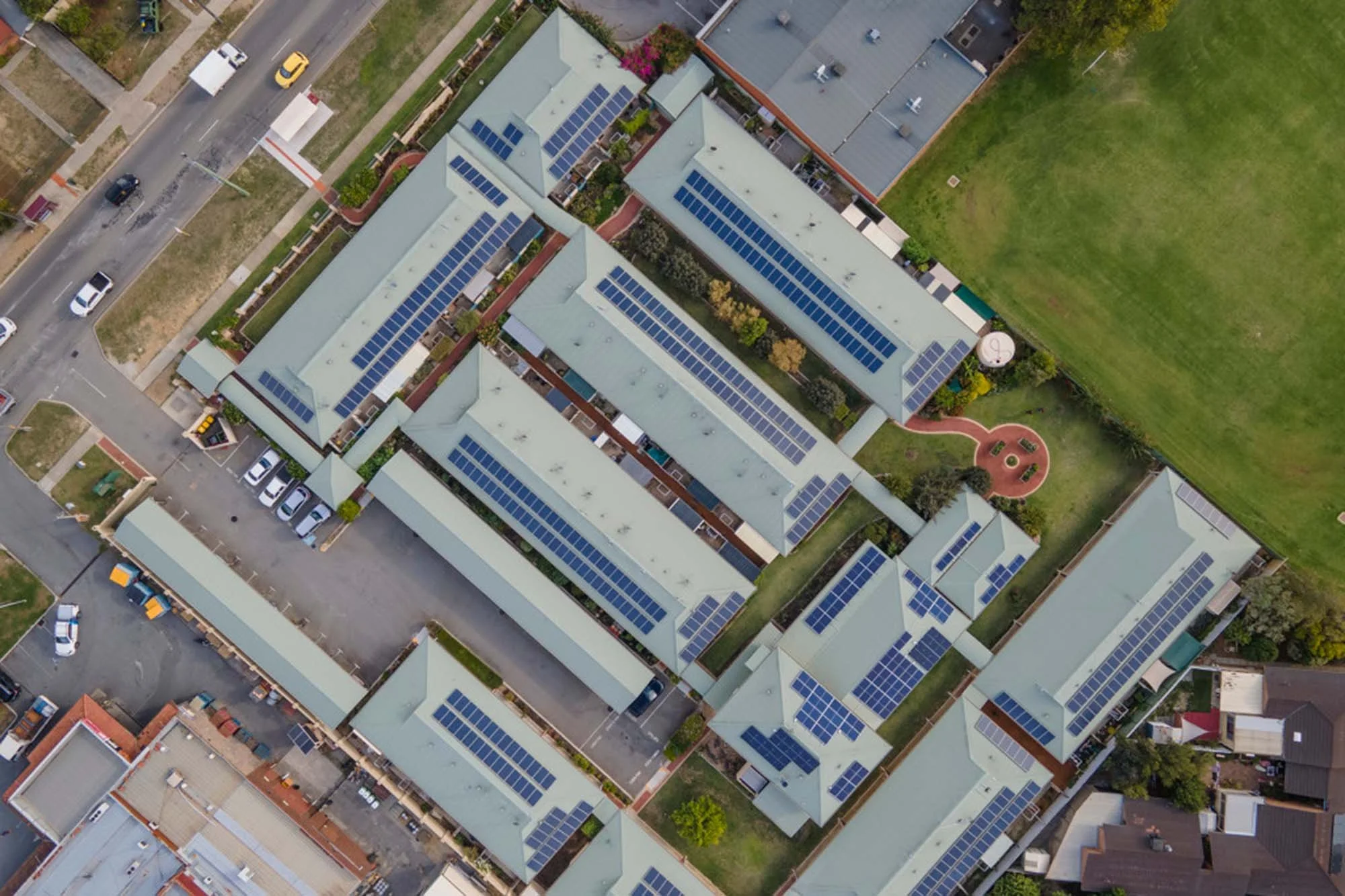 Aerial view of a residential complex with multiple buildings covered in solar panels, surrounded by parking lots and green fields.