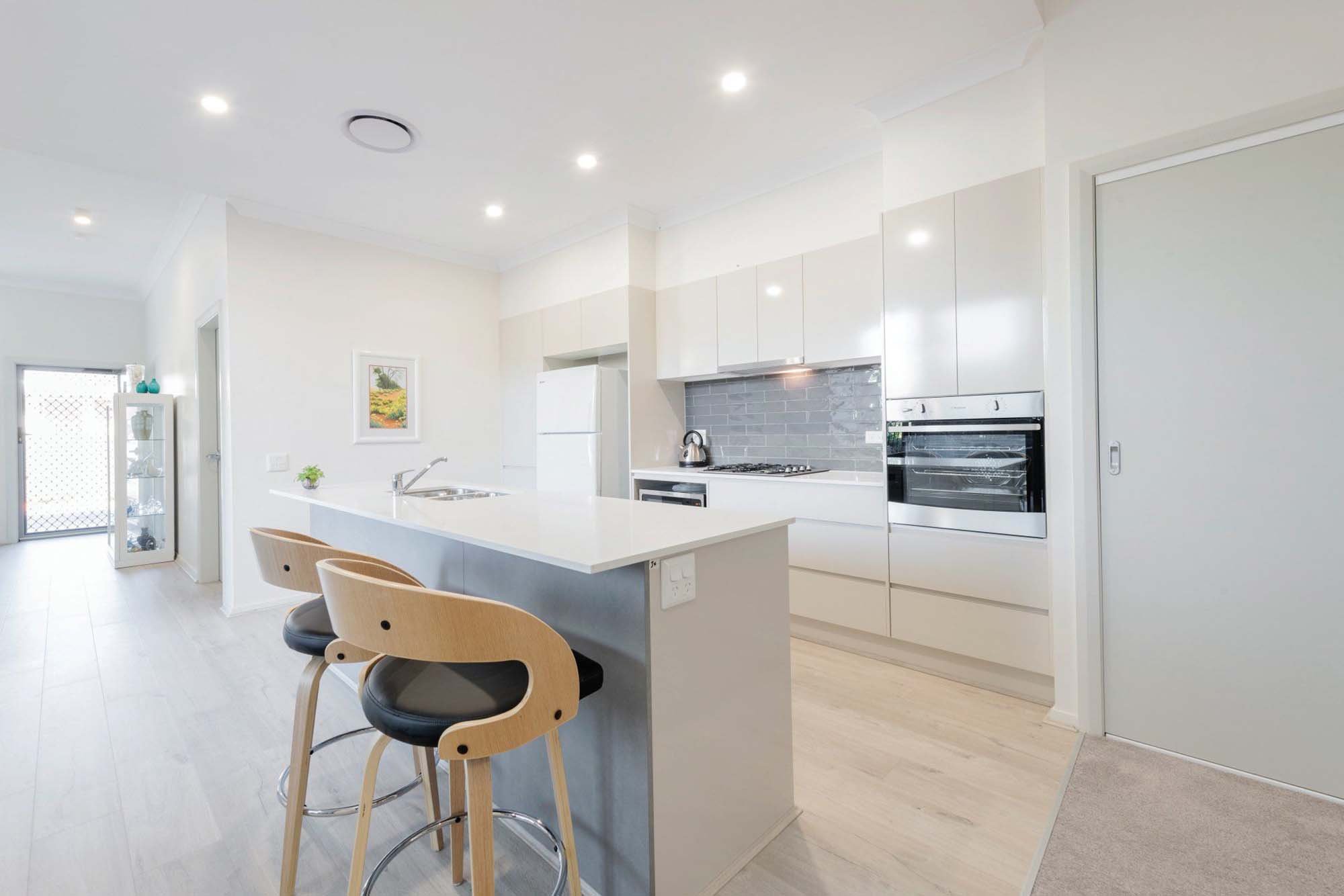 Bright, modern kitchen with white cabinetry, a central island with seating, stainless steel appliances, gray backsplash, and a sliding door leading outside.
