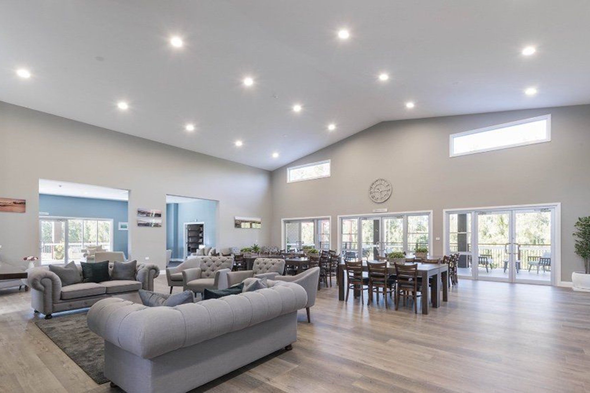 Spacious living and dining area with large windows, light-colored sofas, wooden dining tables, and a high ceiling with recessed lighting.