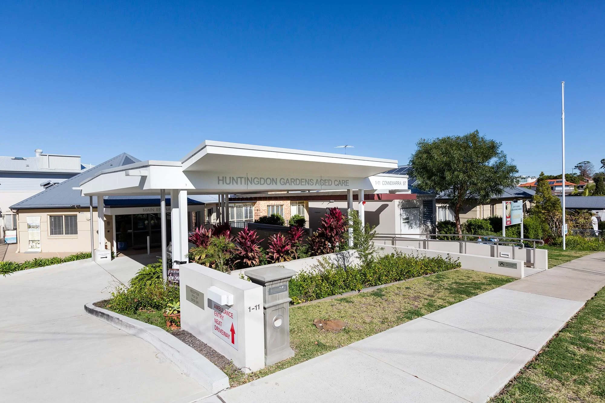 Entrance to Huntingdon Gardens Aged Care facility with a sign, greenery, clear sidewalks, and a blue sky.