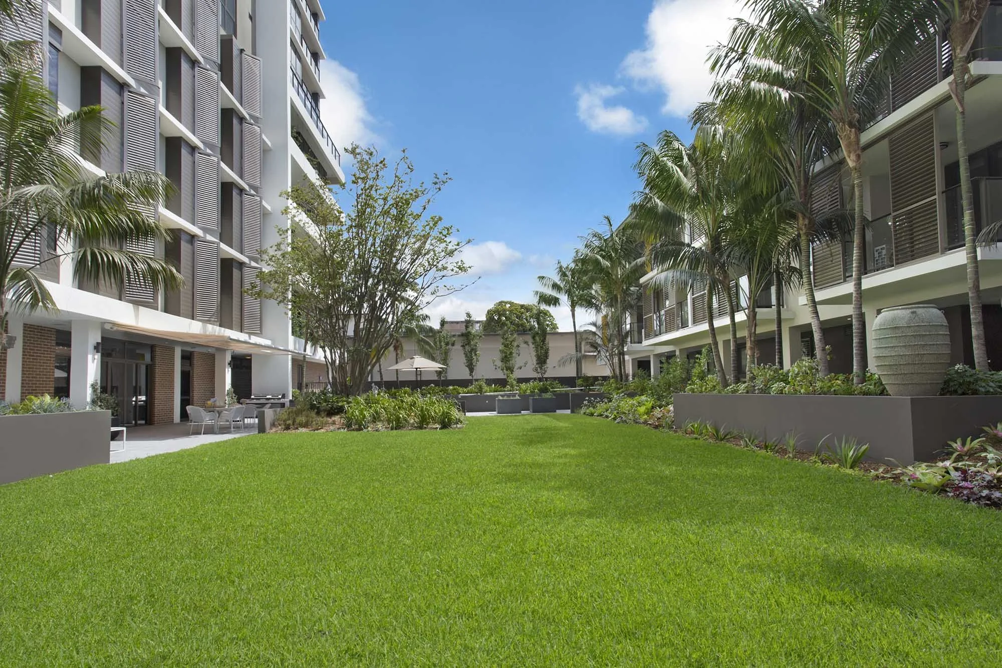A landscaped courtyard in an urban apartment complex with green grass, tropical trees, and outdoor seating areas.