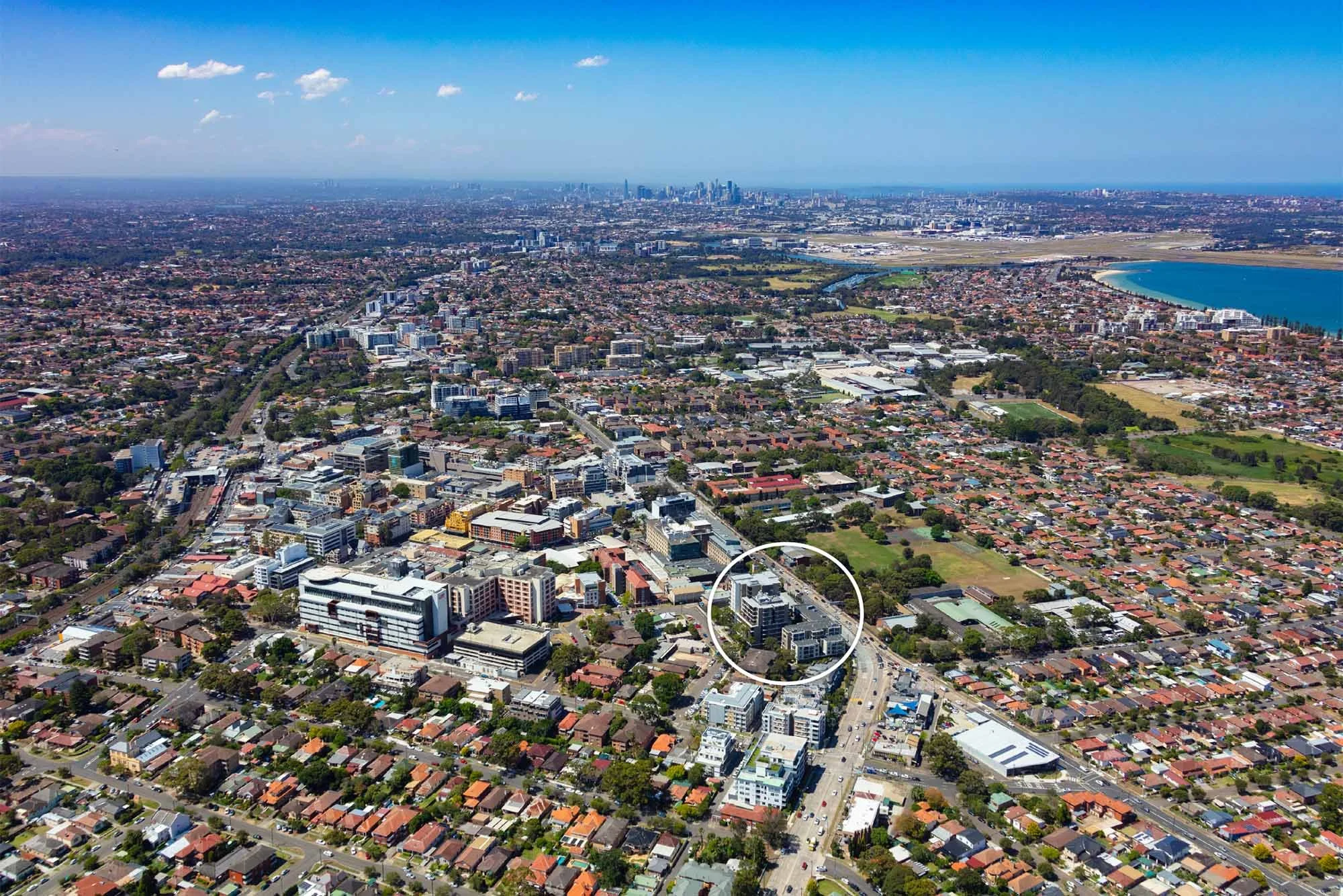 Aerial view of a cityscape showing a mix of high-rise and residential buildings, streets, parks, and a lake in the background, under a partly cloudy sky.