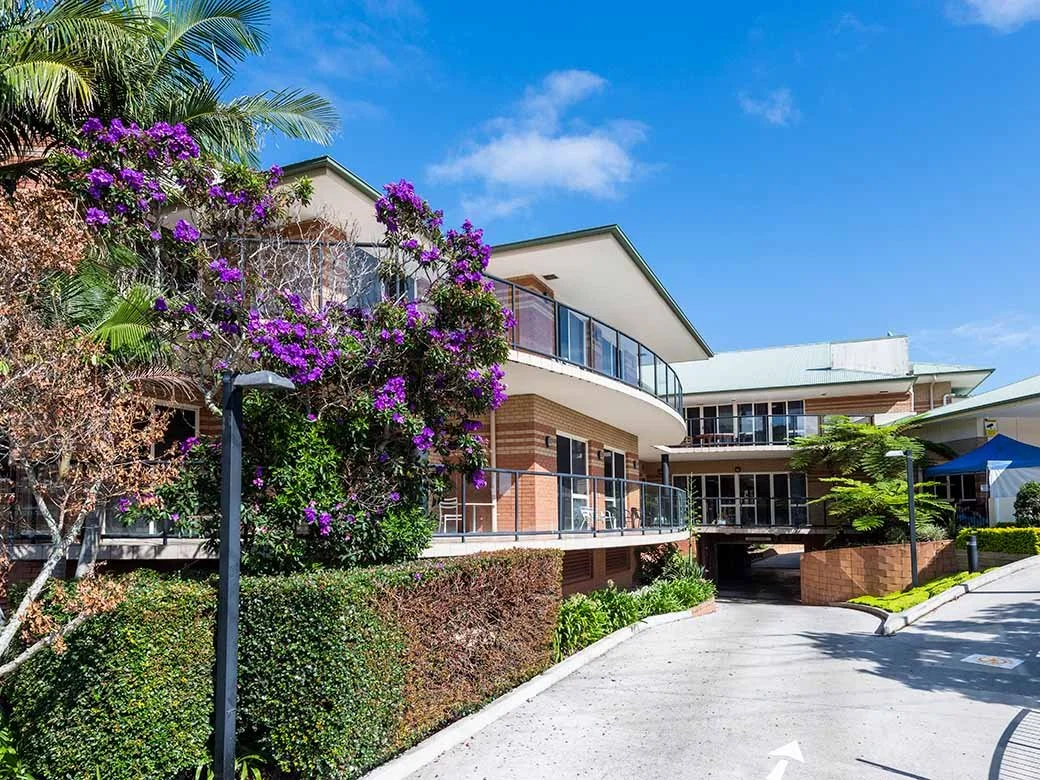 Modern residential building with balconies, lush greenery, and flowering plants outside under a blue sky.