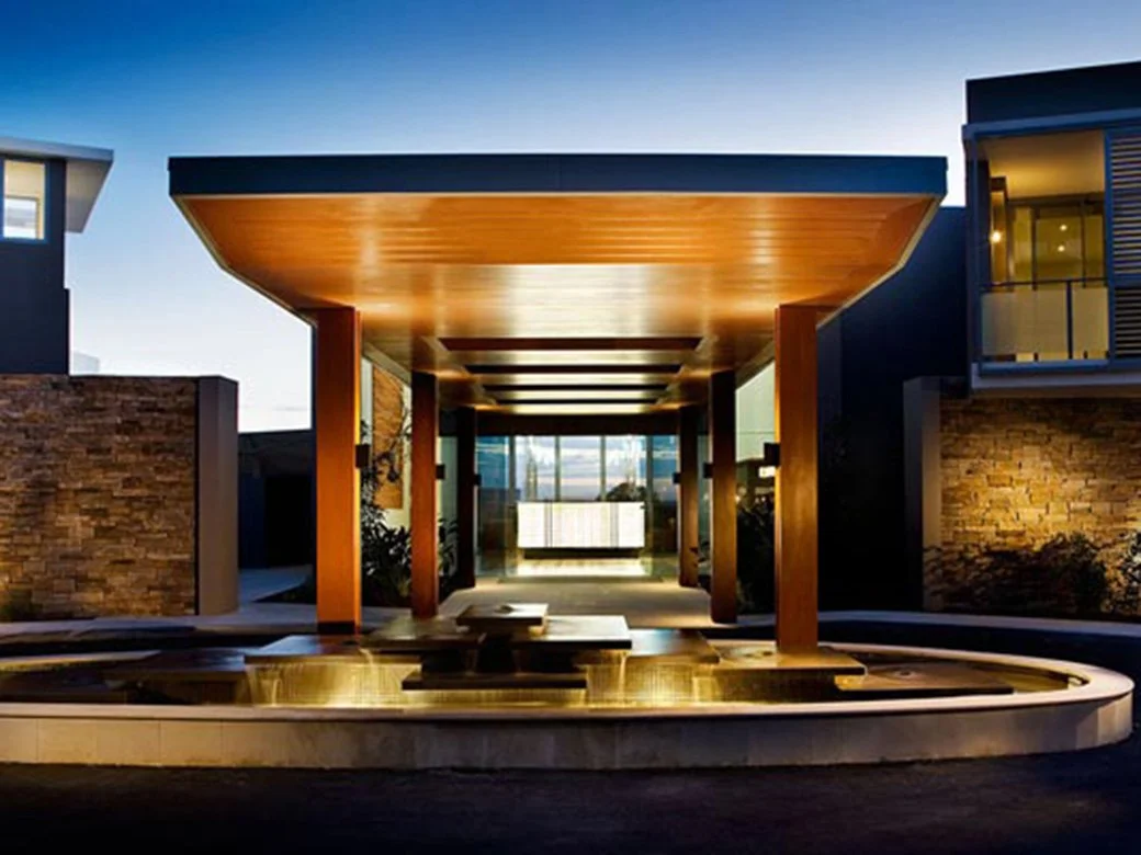 Modern building entrance with a wooden canopy, stone walls, and a fountain in the foreground, illuminated during dusk.