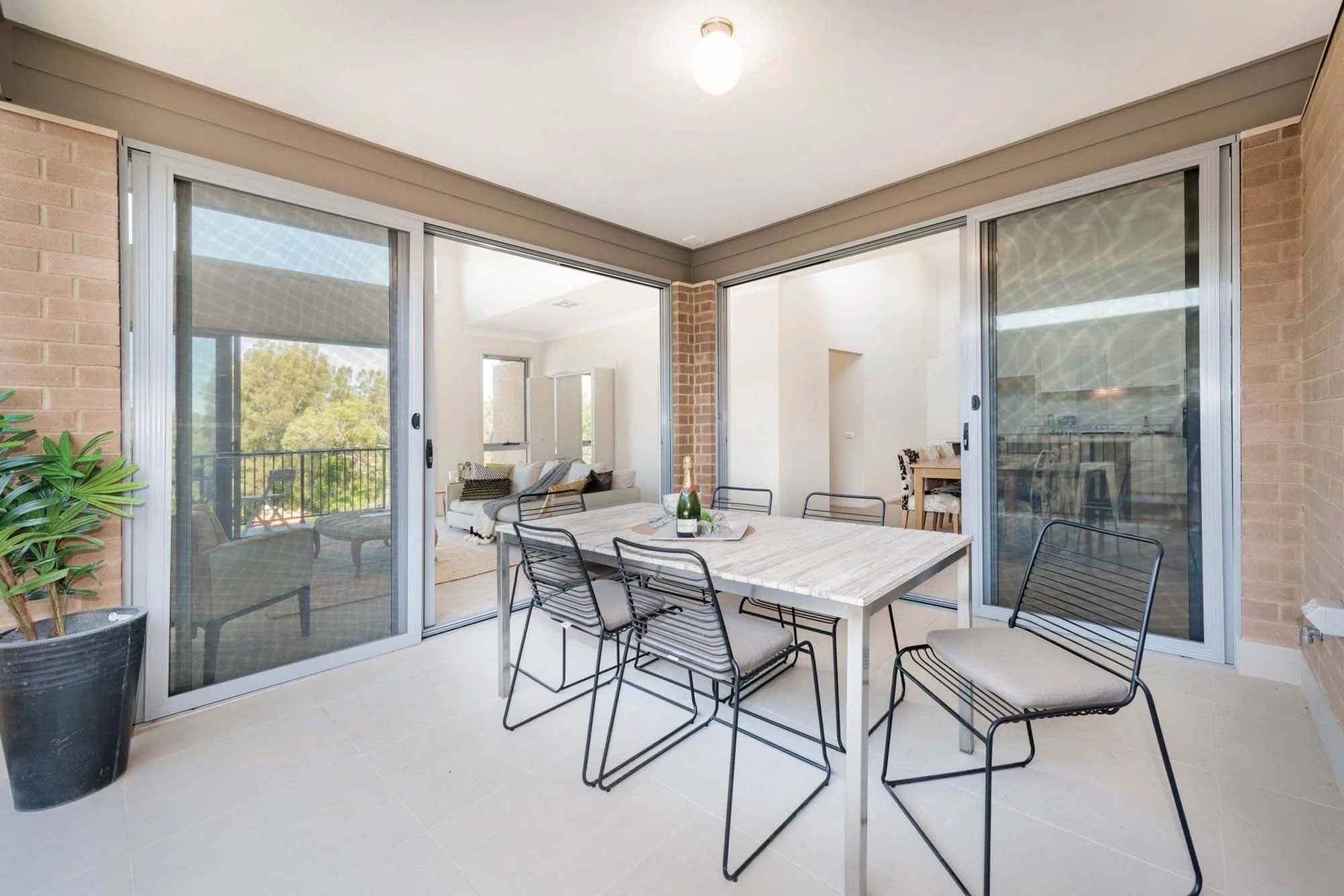 An outdoor patio with glass sliding doors, a dining table with eight metal chairs, a bottle of champagne, and a living room visible in the background.