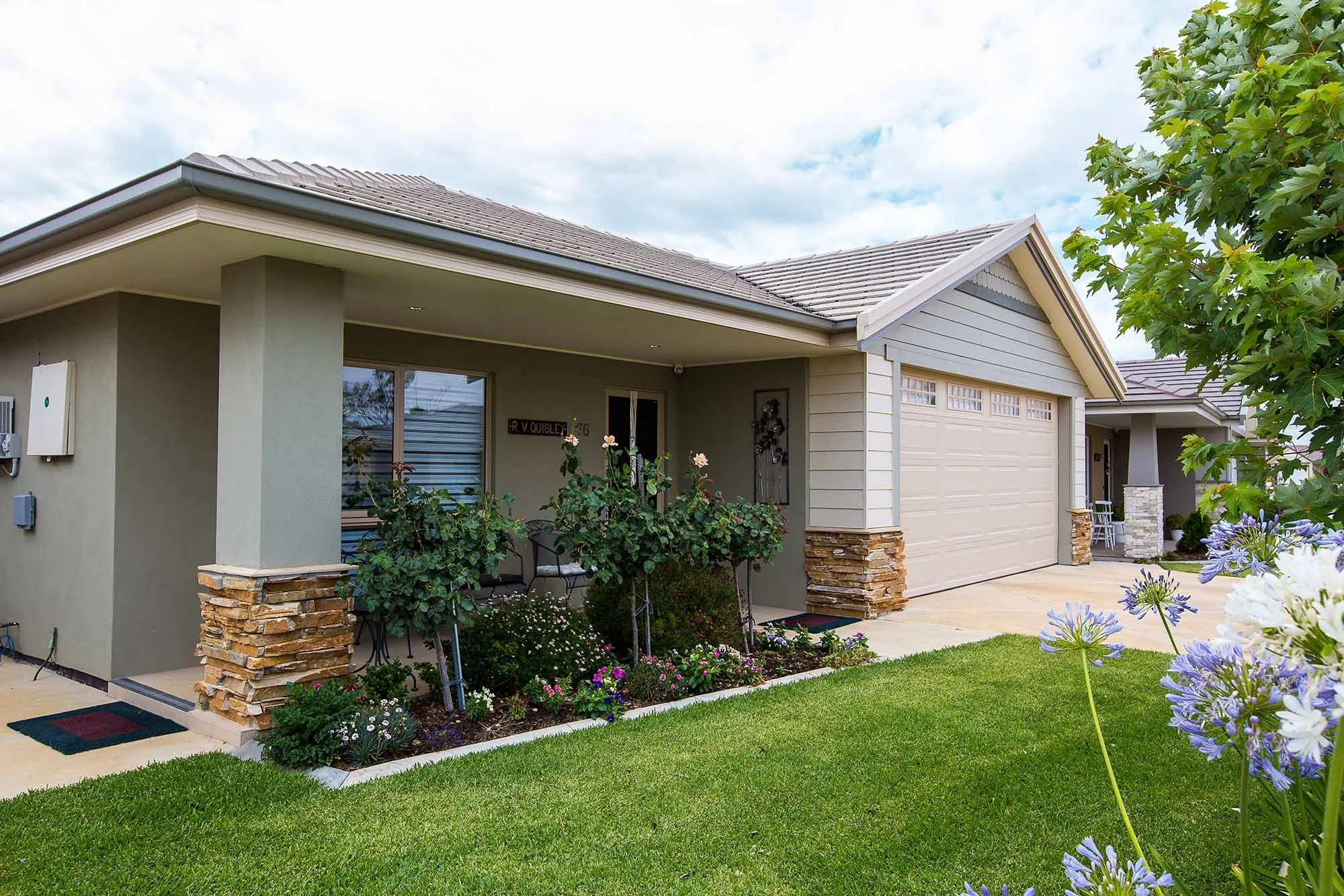 Front view of a modern house with a neatly maintained lawn, flowering plants, and a garage. The house has a covered porch, stone accents at the base of the columns, and a tiled roof.