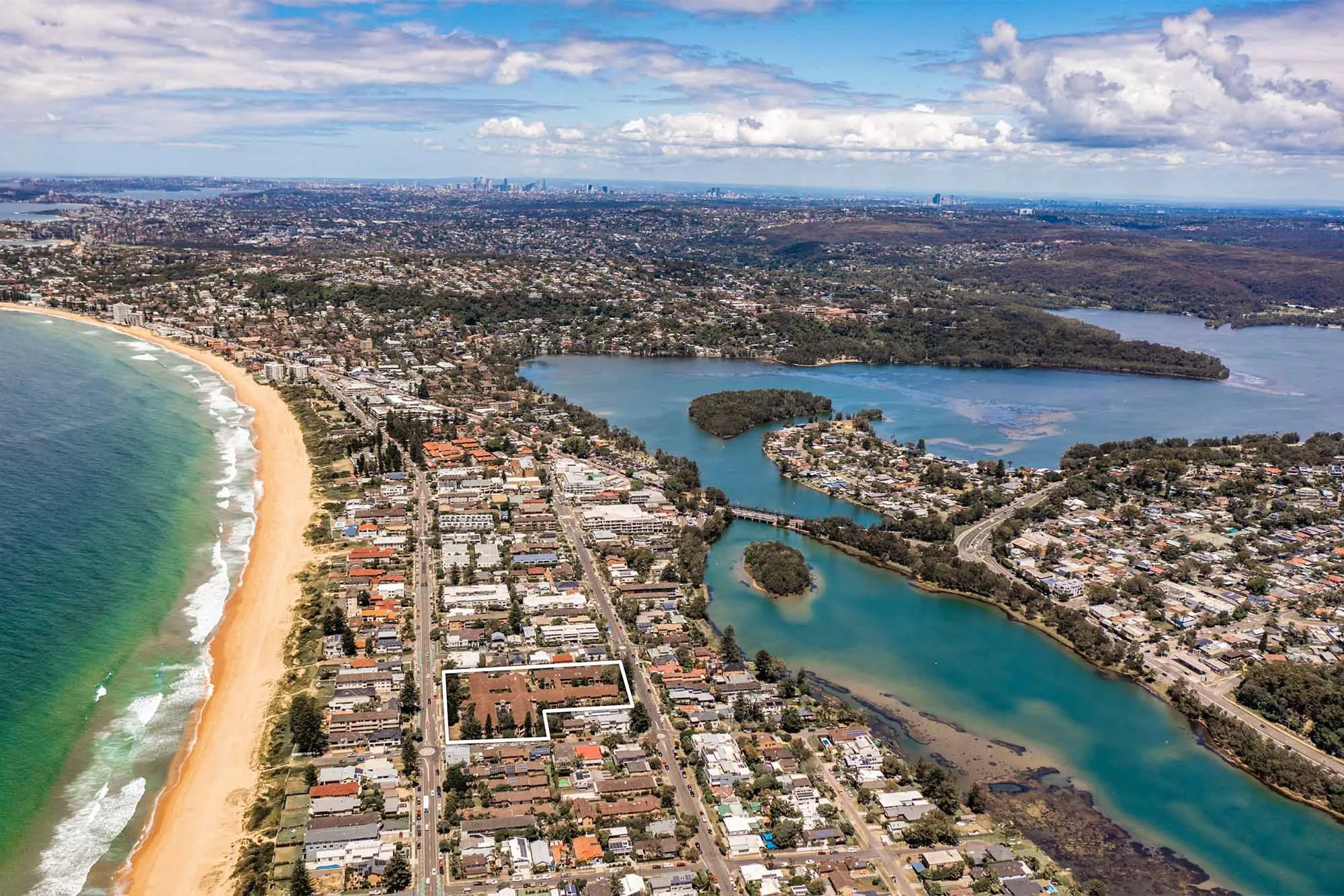 Aerial view of a coastal city with a sandy beach on the left, and a river system with small islands and residential neighborhoods on the right, under a partly cloudy sky.