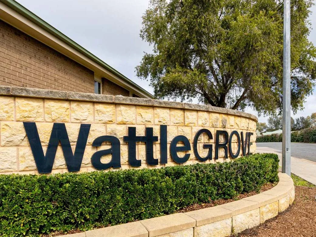 Sign that reads 'WattleGROVE' in front of a brick and stone wall, next to a sidewalk, with a tree and streetlight in the background.