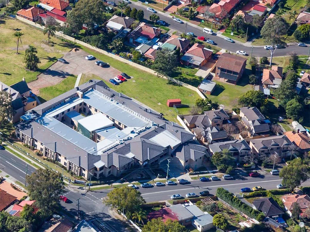 Aerial view of a large U-shaped apartment complex surrounded by houses, parking lots, and green spaces in a suburban neighborhood.