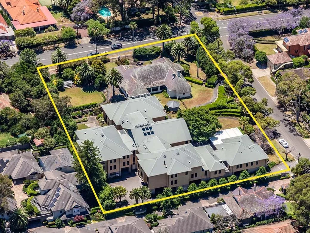 Aerial view of a large residential property outlined in yellow, with multiple buildings, lush green trees, gardens, and surrounding neighborhood houses.