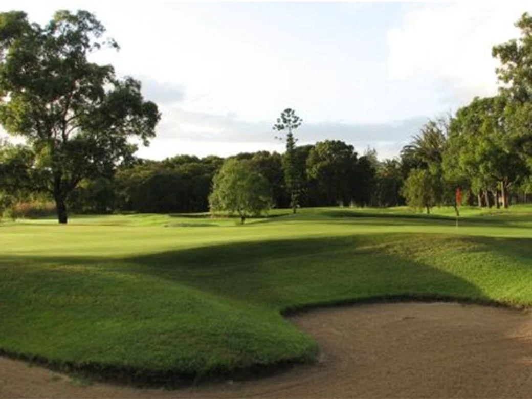 A golf course with well-maintained green grass, trees, and sand bunker, under a partly cloudy sky.