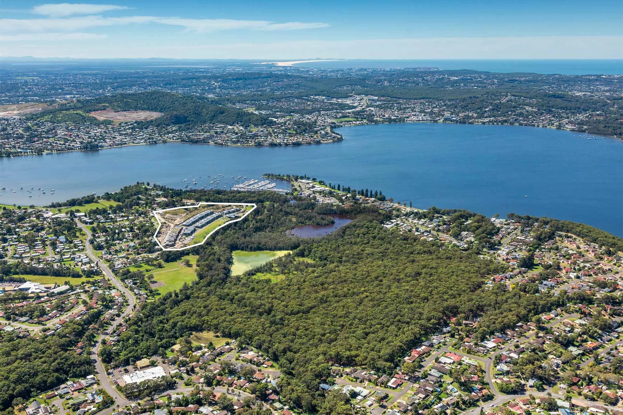 Aerial view of a large lake surrounded by green wooded areas and residential neighborhoods, with a boat marina along the shore and distant cityscape in the background.